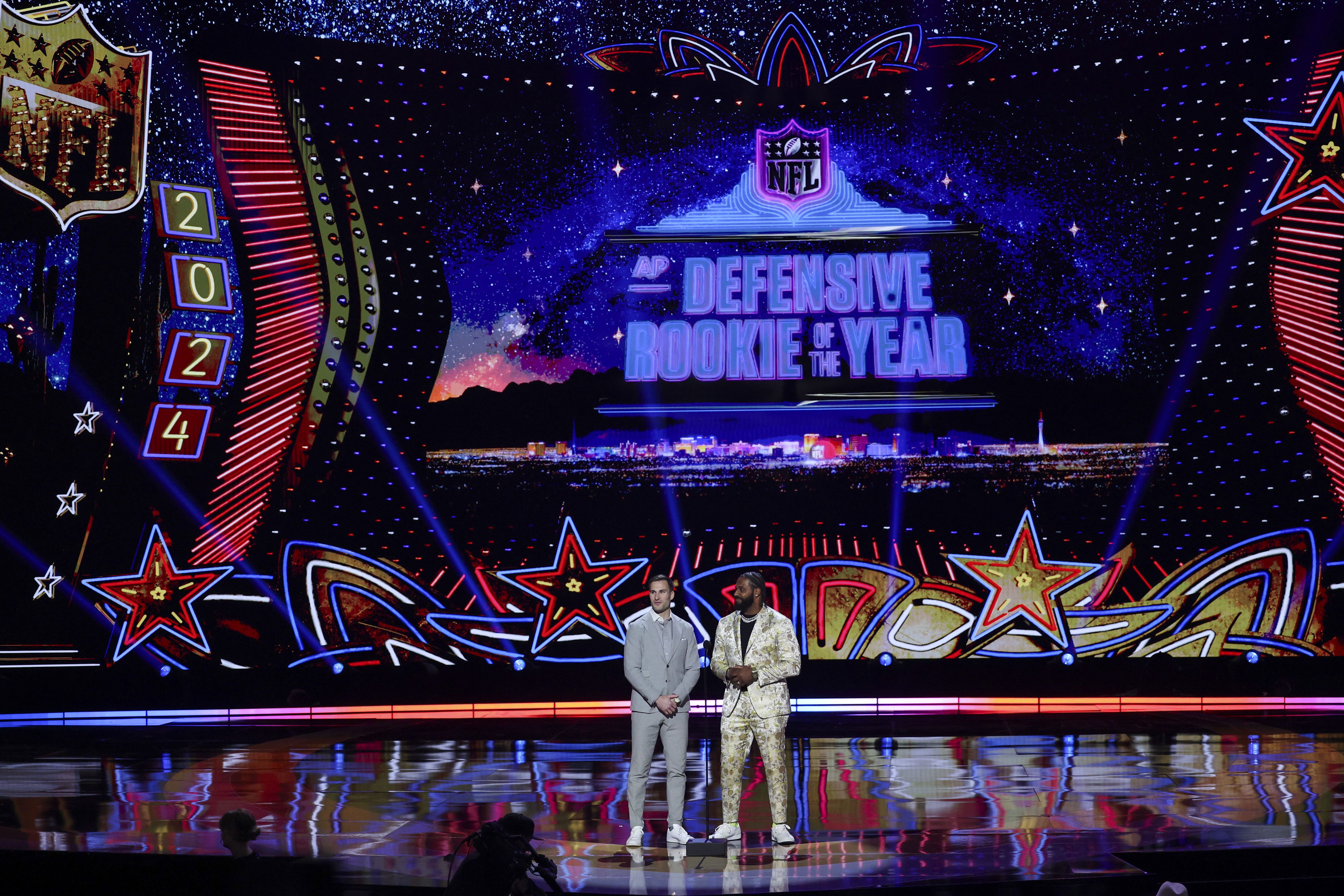 Two men stand in front of a microphone on an empty stage. Behind them is a Las Vegas-themed image that says "Defensive Rookie of the Year."