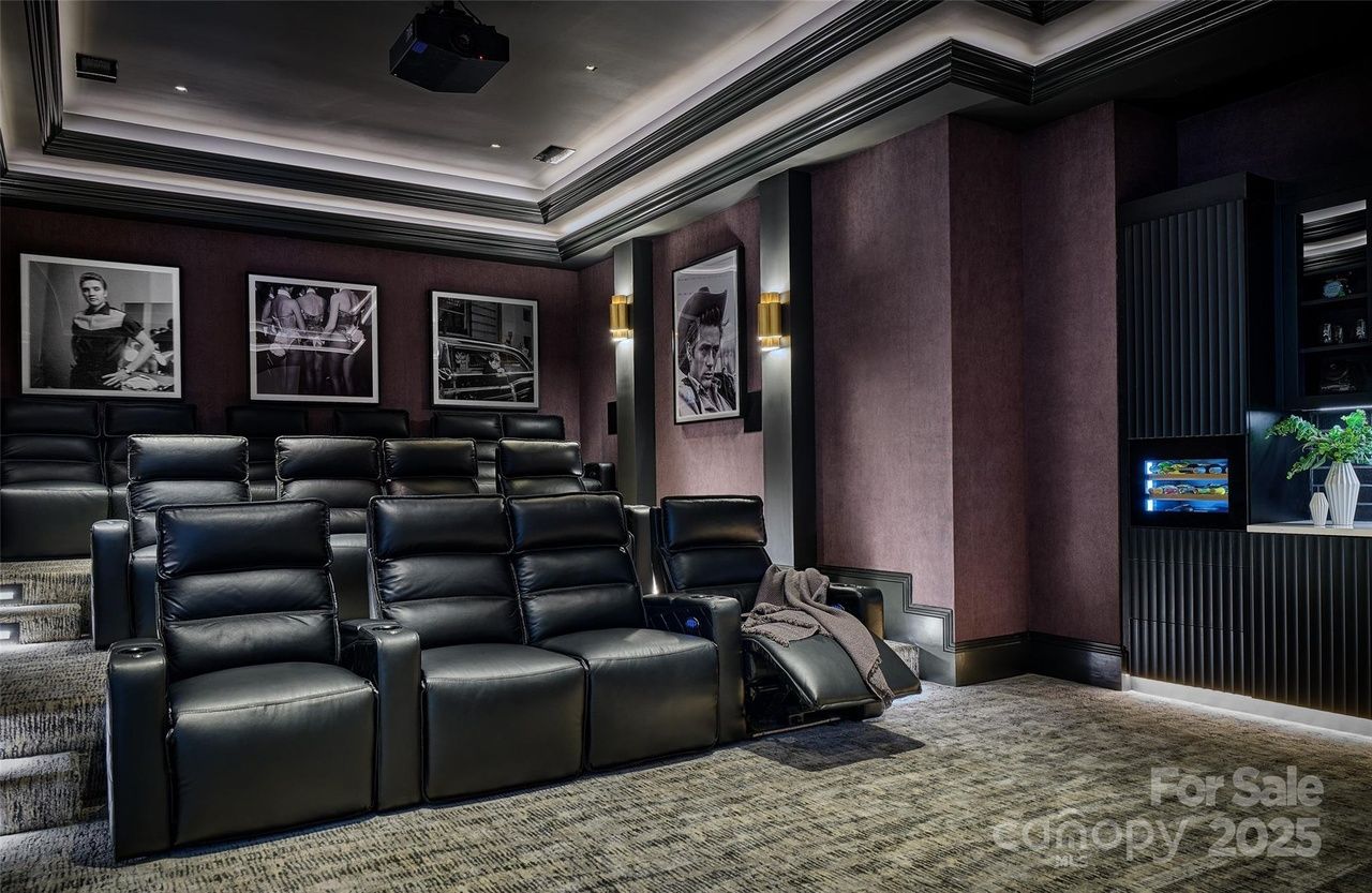 Luxurious home theater with black leather recliners, purple walls adorned with black-and-white movie posters, gray carpeted floor, modern lighting, and a cabinet with a glowing wine cooler.