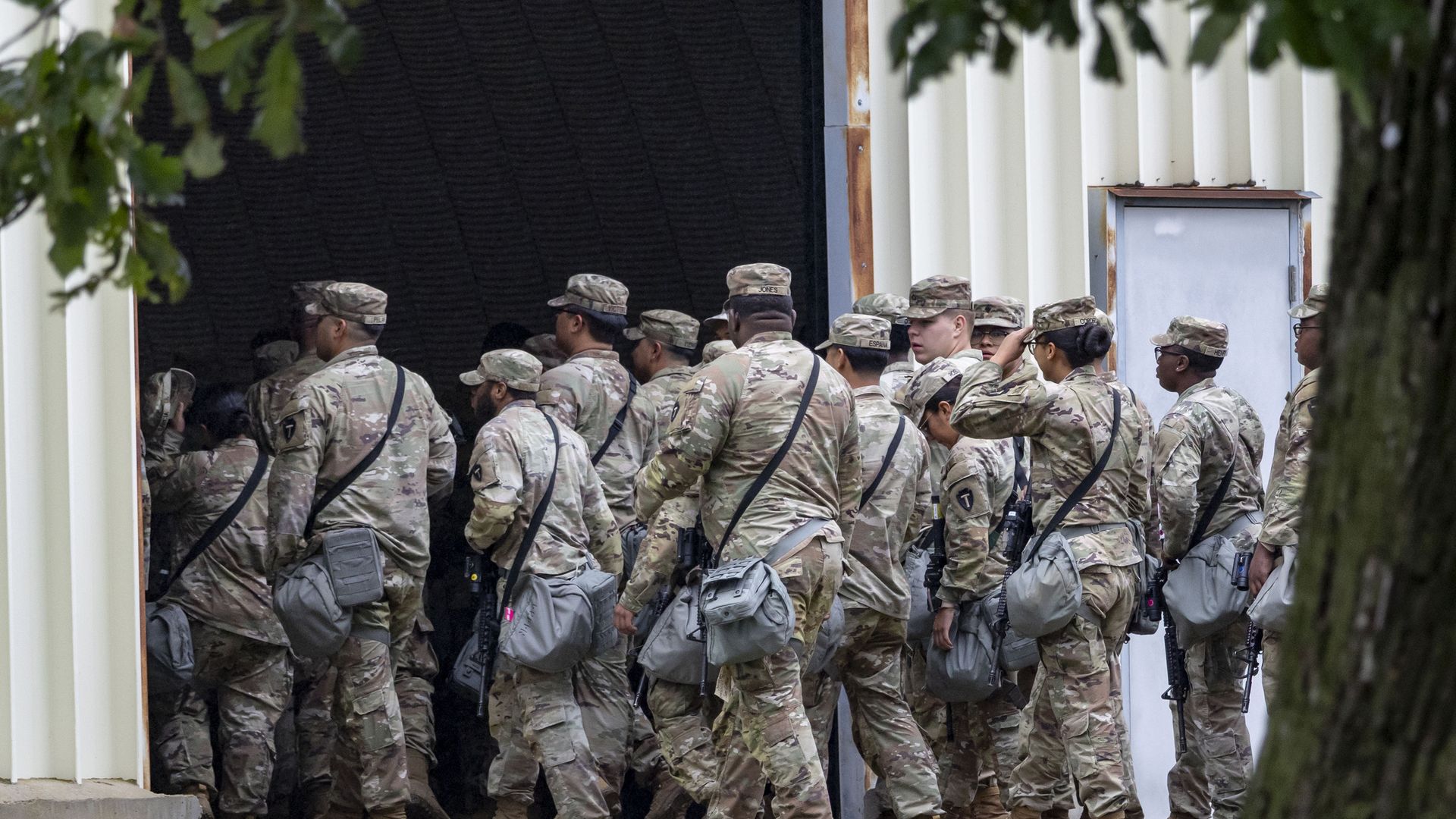 A group of soldiers in camo military uniforms walk from outside into a large doorway.