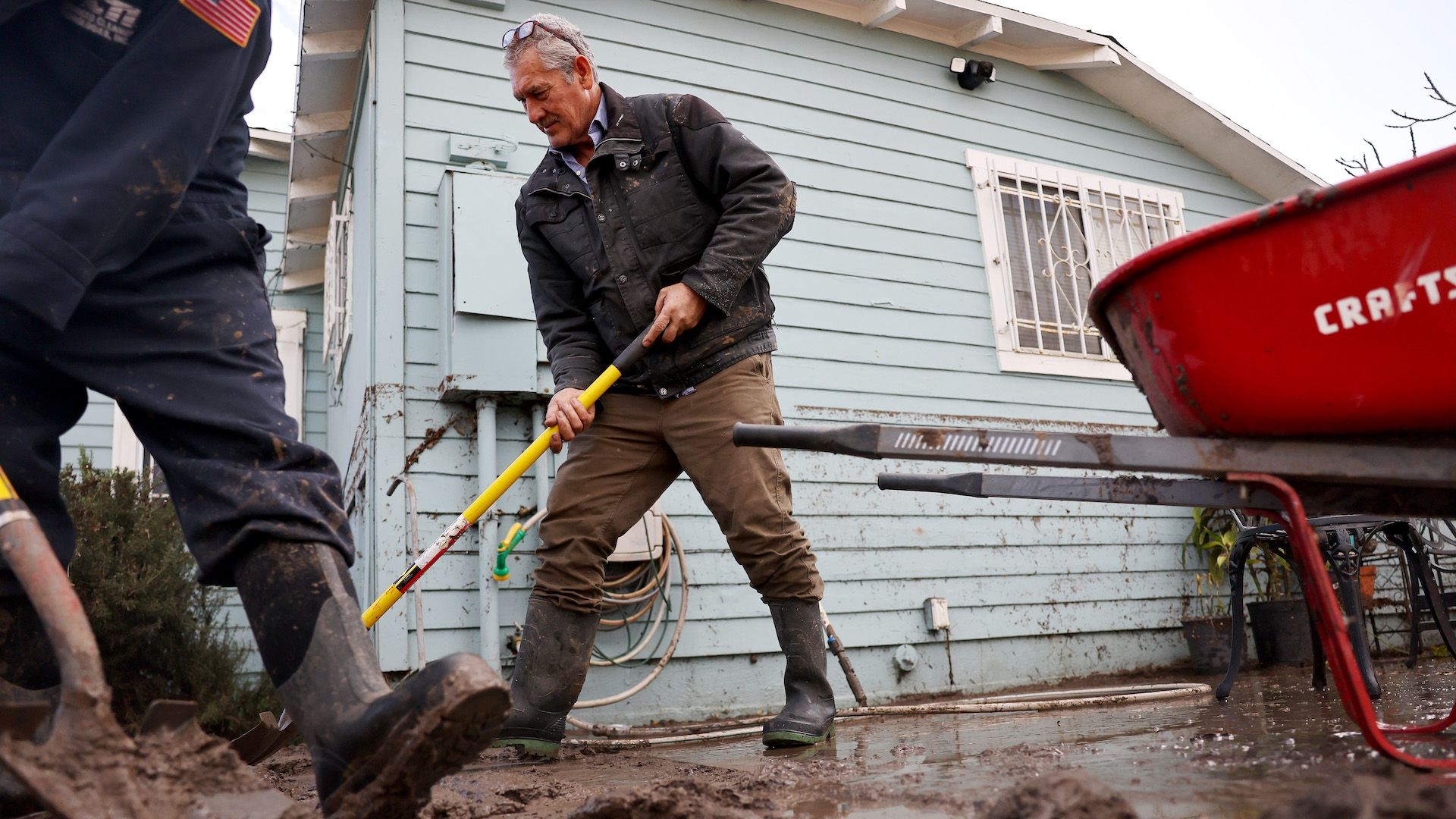Two men man shovel debris and mud into a red wheelbarrow outside a flooded home.