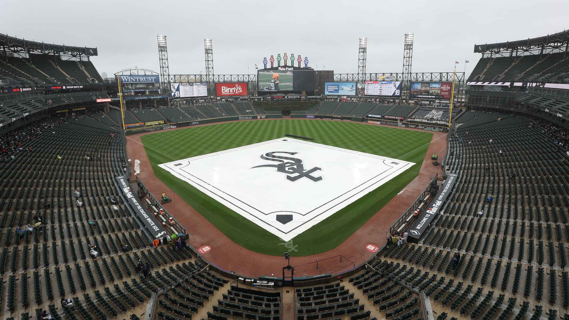 Wide, mostly empty baseball stadium on a gray, overcast day; the infield is covered by a large white tarp bearing the Chicago White Sox logo, with tall floodlights and scoreboards behind.