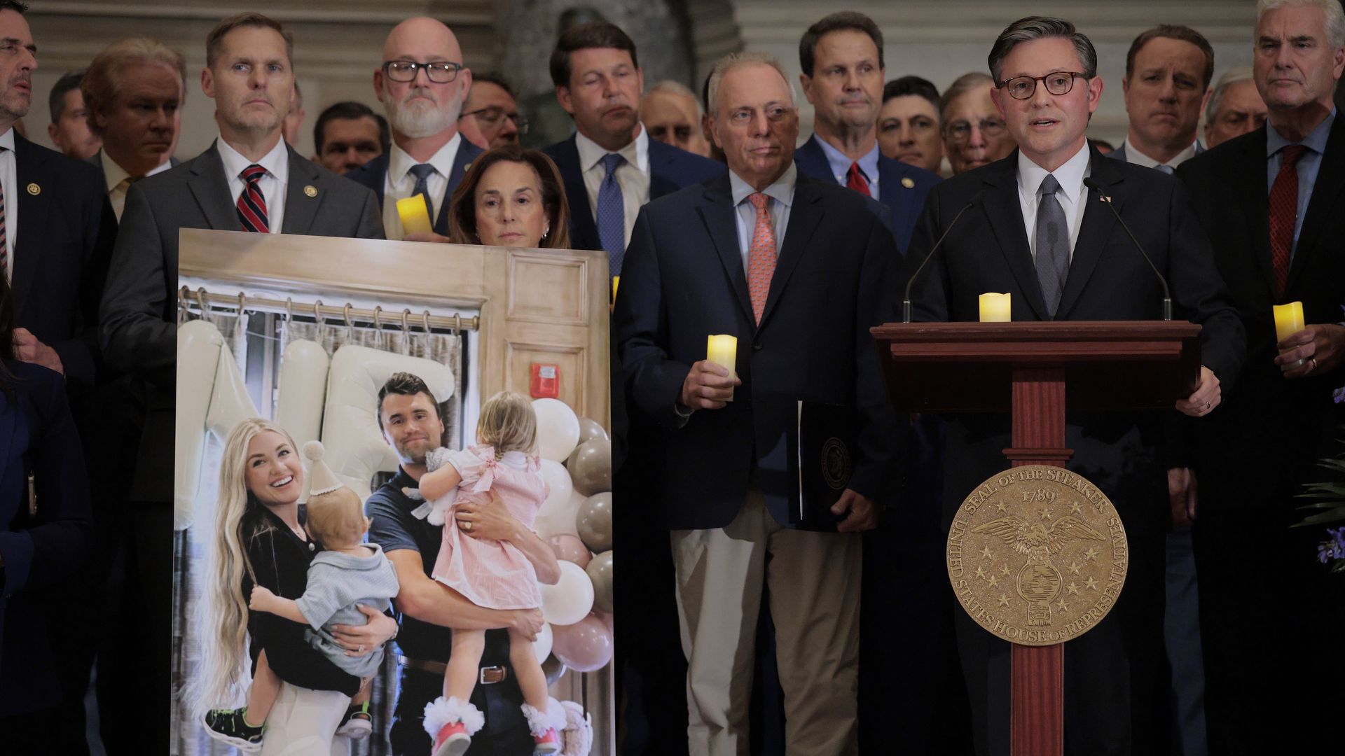 Mike Johnson speaking at a podium with GOP lawmakers behind him and a photo of Charlie Kirk and his family next to him