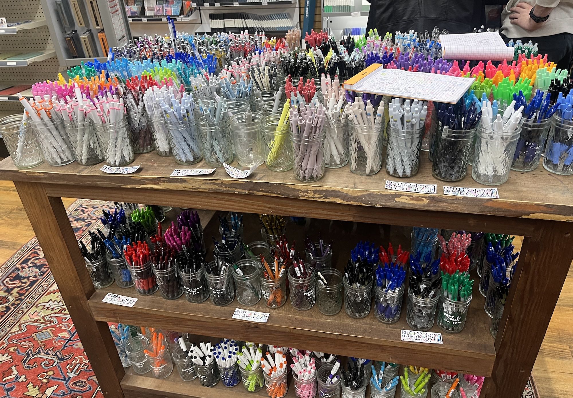 A wooden table with three shelves filled with various colorful pens organized in clear jars, with handwritten price tags indicating $2 and $2.79 in a store setting.