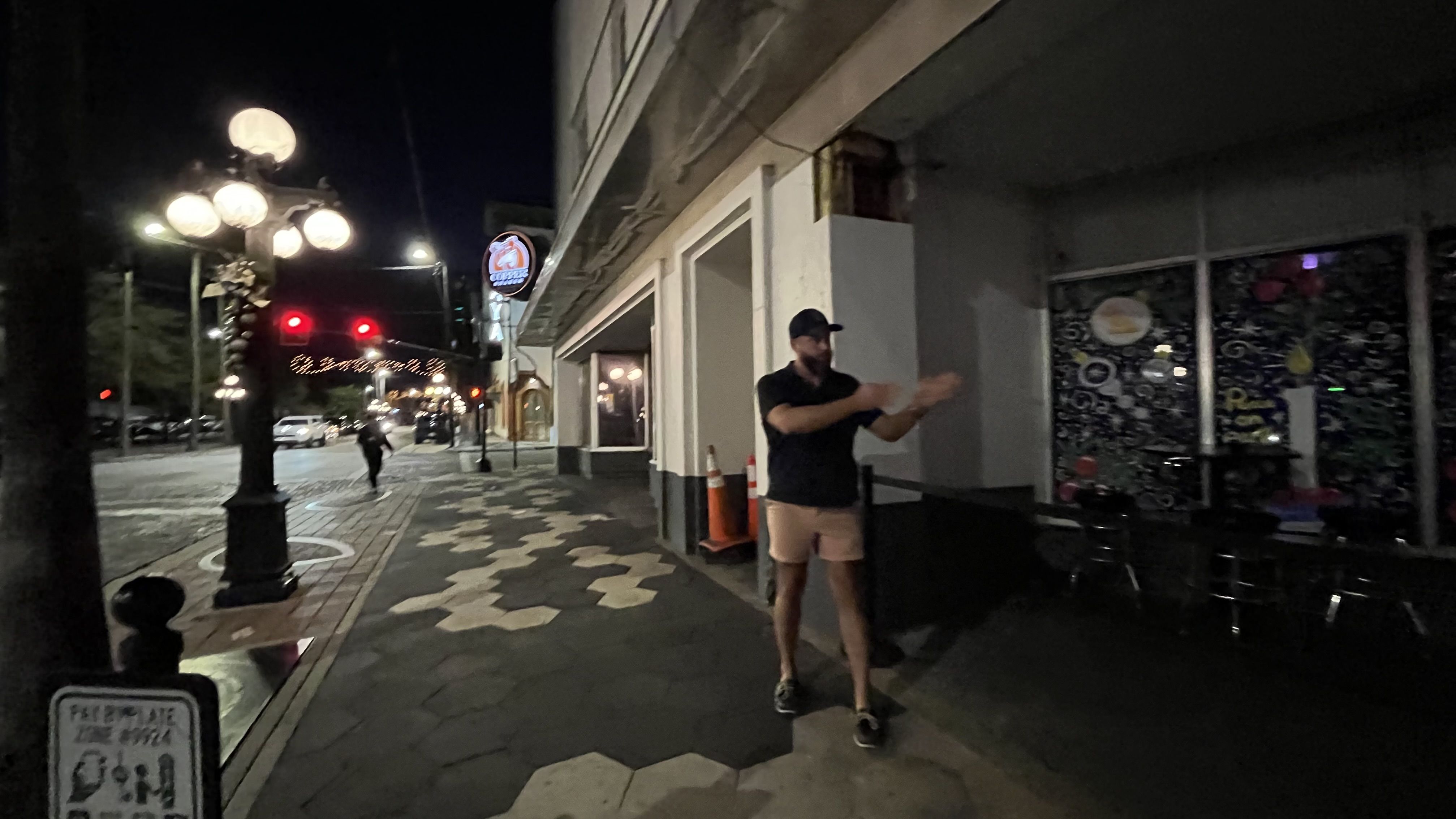 Night street scene with a man in a black shirt and shorts walking on a patterned sidewalk near a building. A large street lamp and traffic lights glow red in the background.