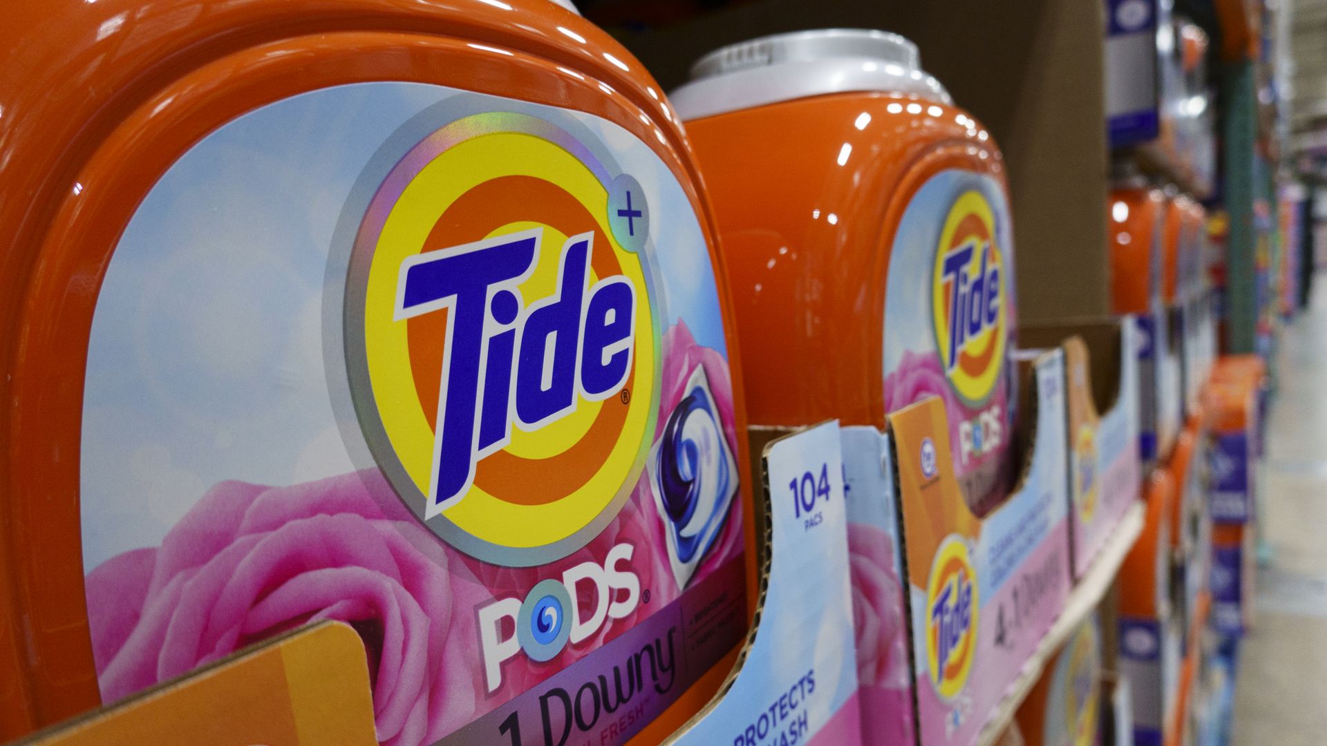 Close-up of orange Tide Pods detergent containers with 4-in-1 Downy April Fresh scent, stacked on store shelves in cardboard packaging showing 104 pods per container.