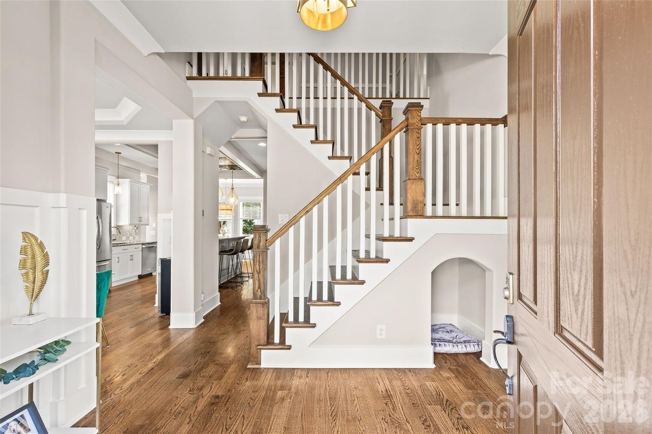 Bright open entry with a two-tone staircase: white spindles, dark wood handrail. Left: white kitchen with stainless fridge; warm wood floors. Under stairs, a small alcove with a cushion.