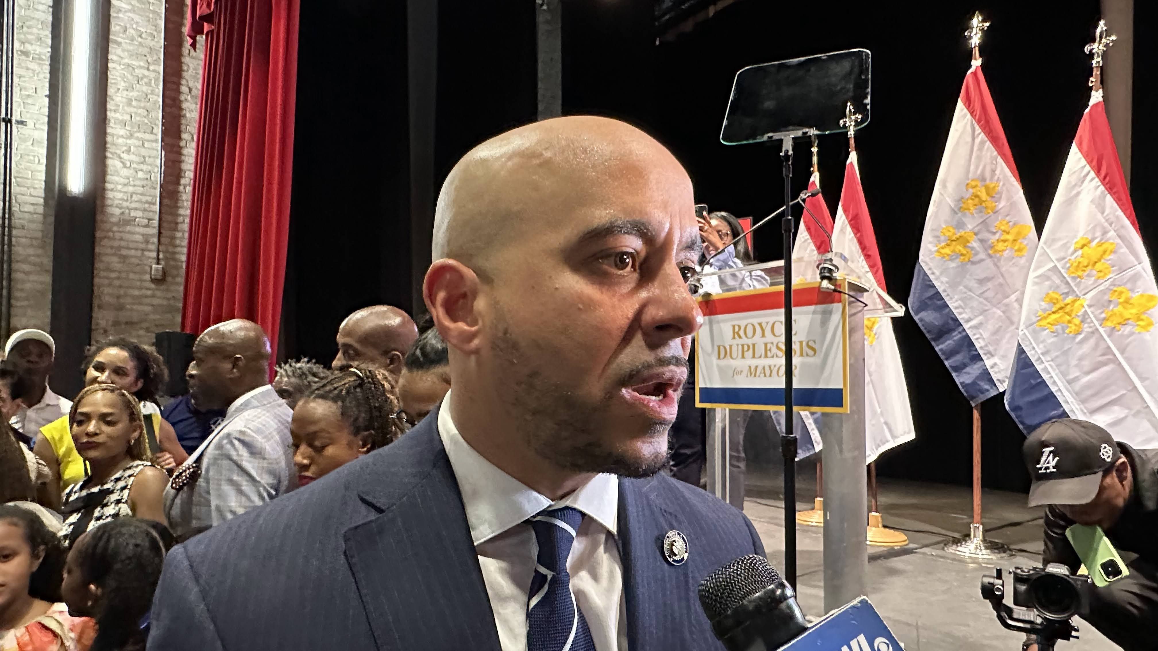 A man in a blue suit and striped tie speaks into a microphone at an indoor event with a crowd and flags in the background, including a sign that reads "Royce Duplessis for Mayor."
