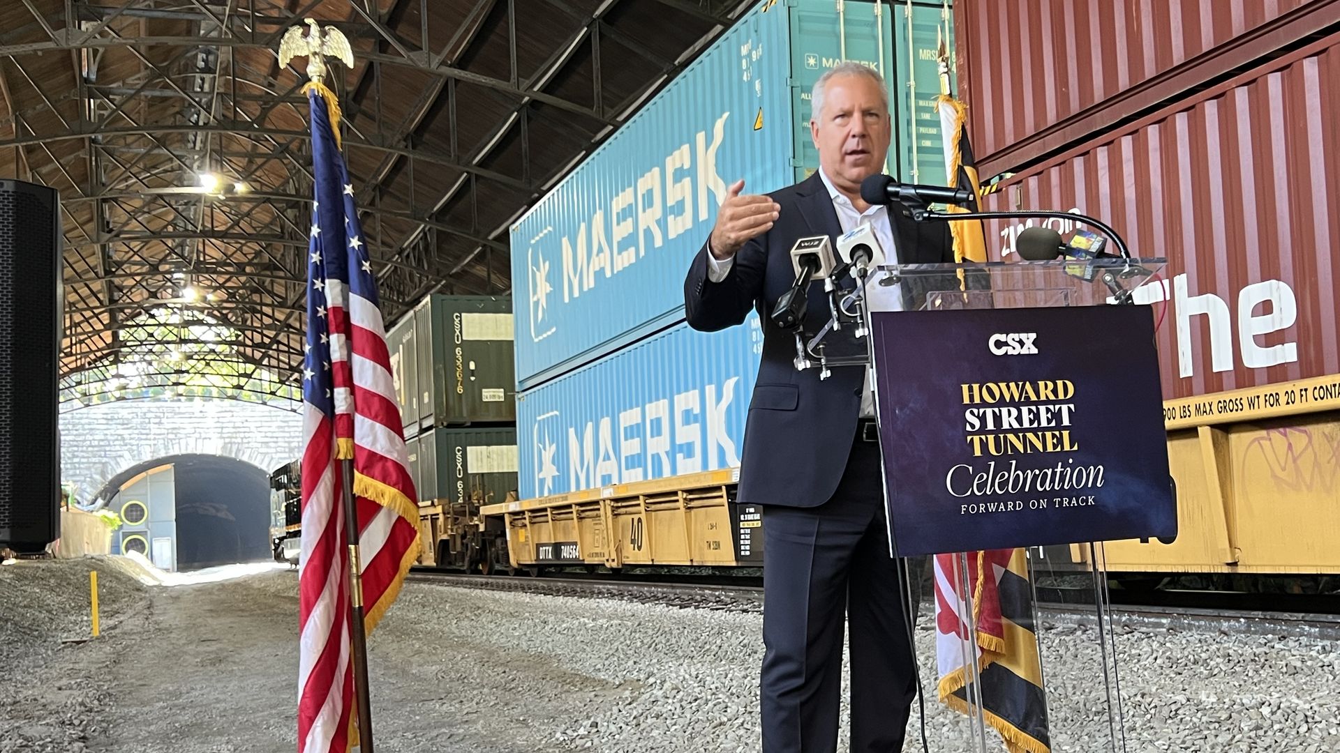 Man in suit speaks at podium with sign reading "HOWARD STREET TUNNEL Celebration" in front of stacked Maersk shipping containers inside a covered train tunnel with an American flag nearby.