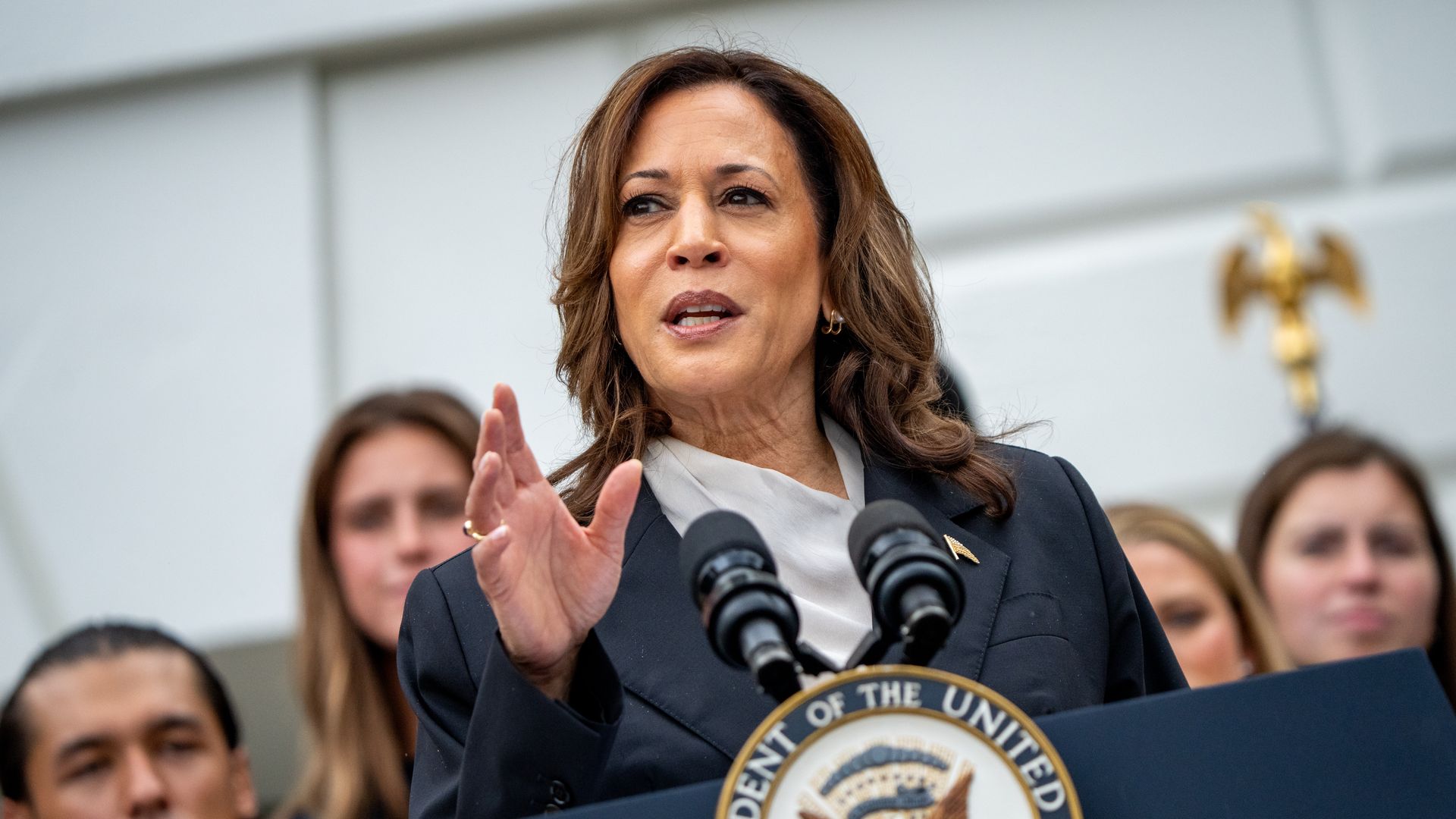 Kamala Harris stands at a podium with an official presidential logo and people behind her as she speaks with her hand raised slightly in front of her.