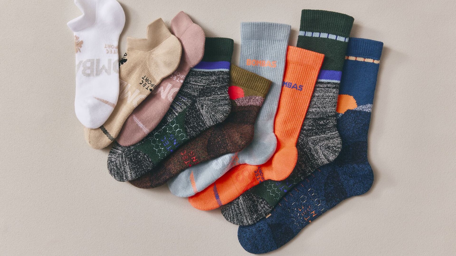 A row of colorful socks fanned out on a beige background, featuring white, beige, pink, green, gray, orange, and blue socks with patterns; several socks show the brand name BOMBAS.