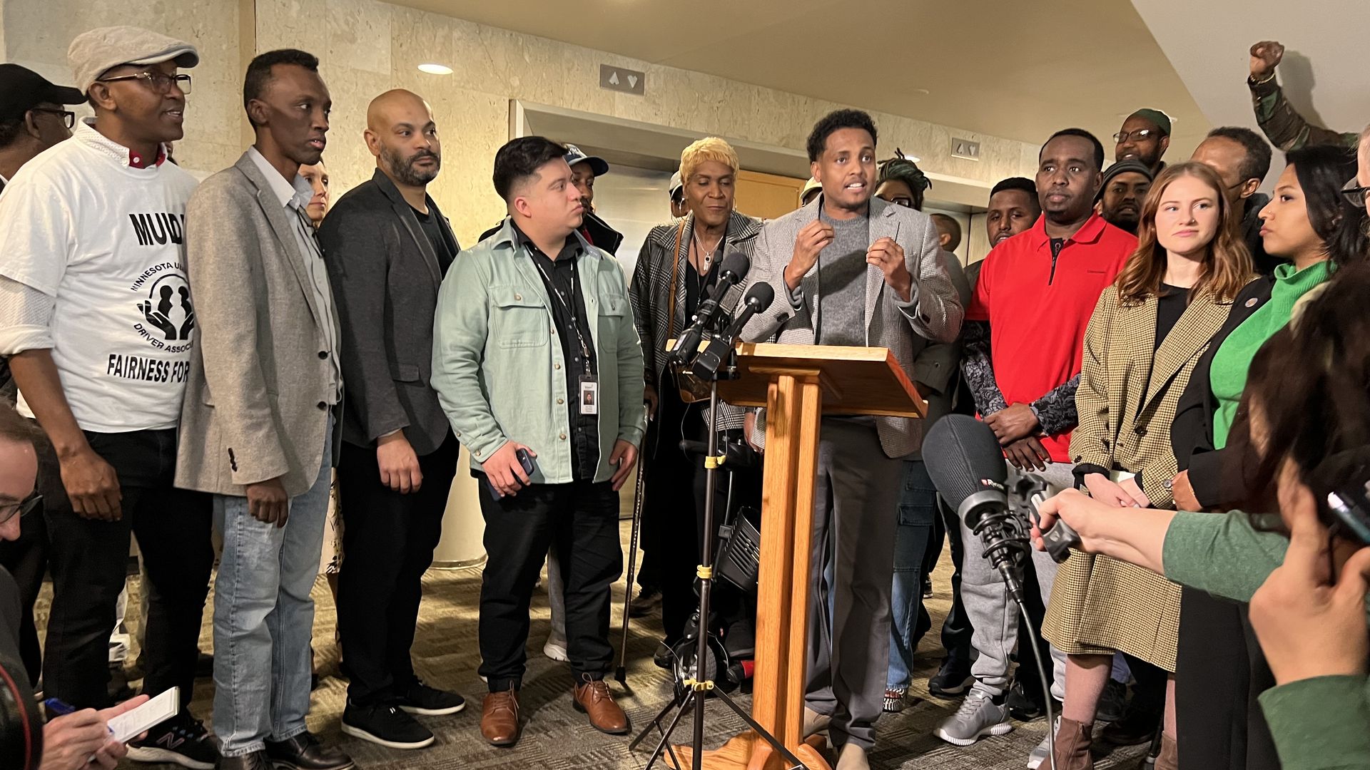 A group of more than two dozen people stand around a podium with microphones. A man with dark skin stands in front of the microphones, speaking and gesturing as others look on.