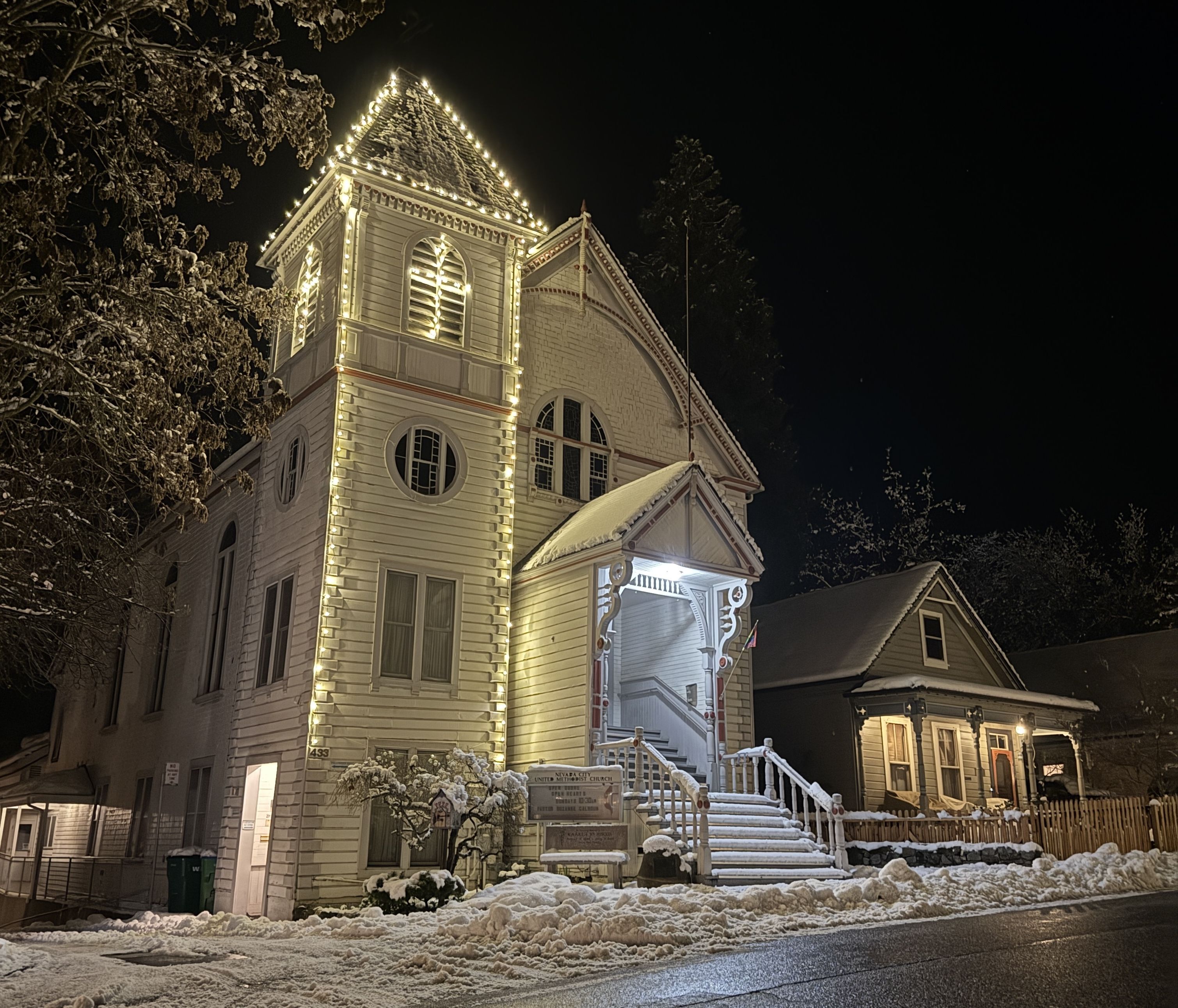 Night view of a snow-covered white church decorated with warm white string lights outlining the tower and roof, with adjacent houses also covered in snow along a snowy street.