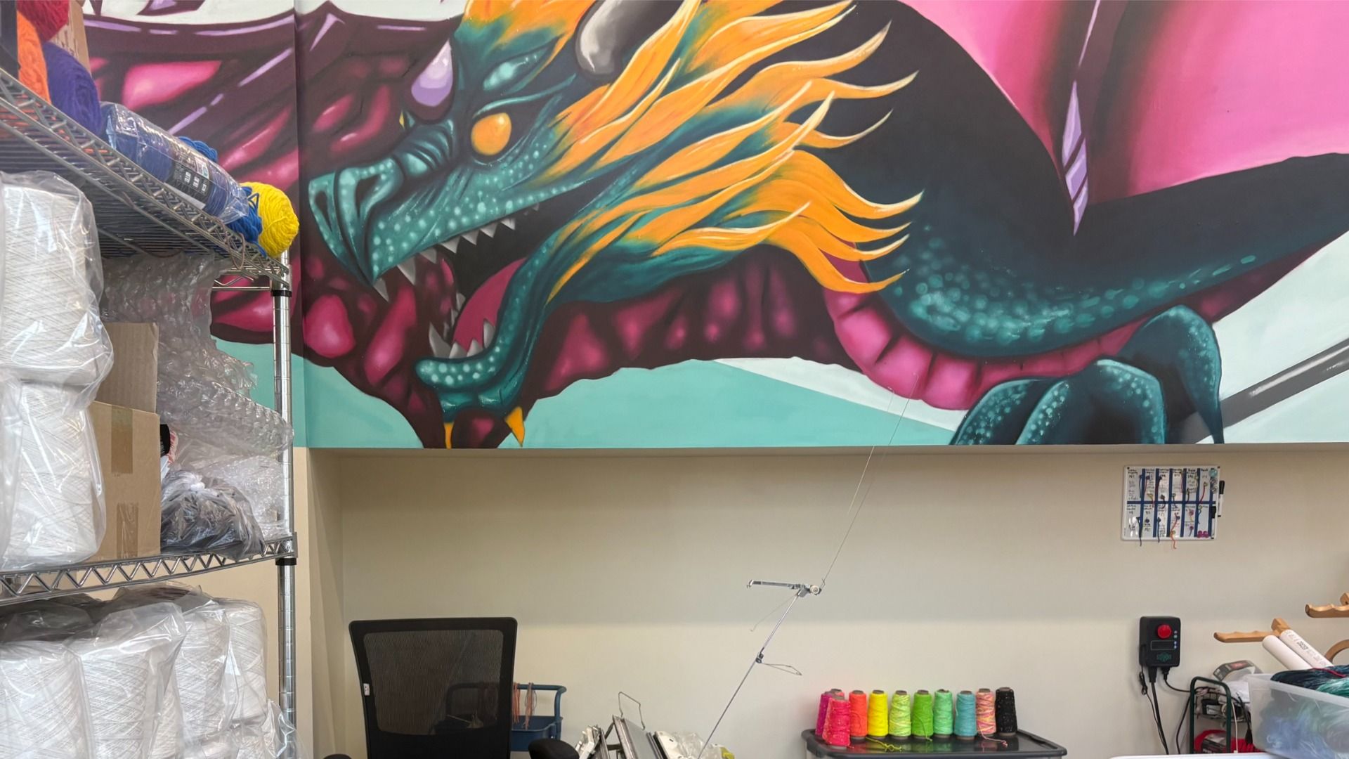 Bright mural of a teal dragon with orange mane on a workshop wall, left shelves stacked with fabric and yarn, and a row of colorful thread spools on a table.