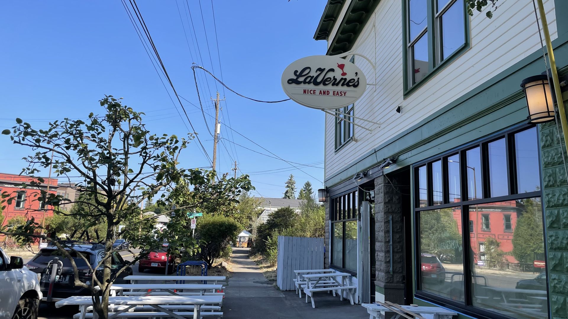 Outdoor scene of a white and green building with a sign reading LaVerne's Nice and Easy, white picnic tables outside, trees, parked cars, and a clear blue sky.