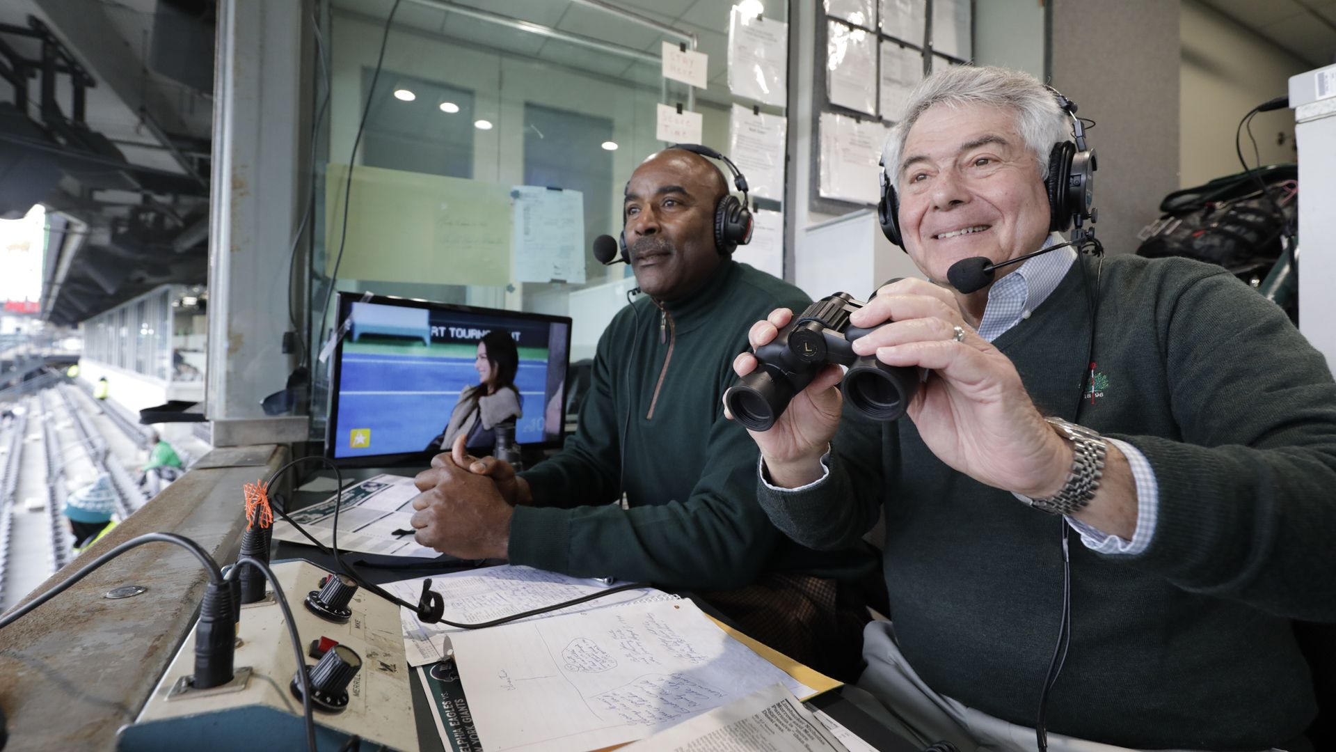 Two men in a broadcast booth wearing headphones with microphones; one holds binoculars smiling, with papers and equipment on the desk, stadium seating visible outside.