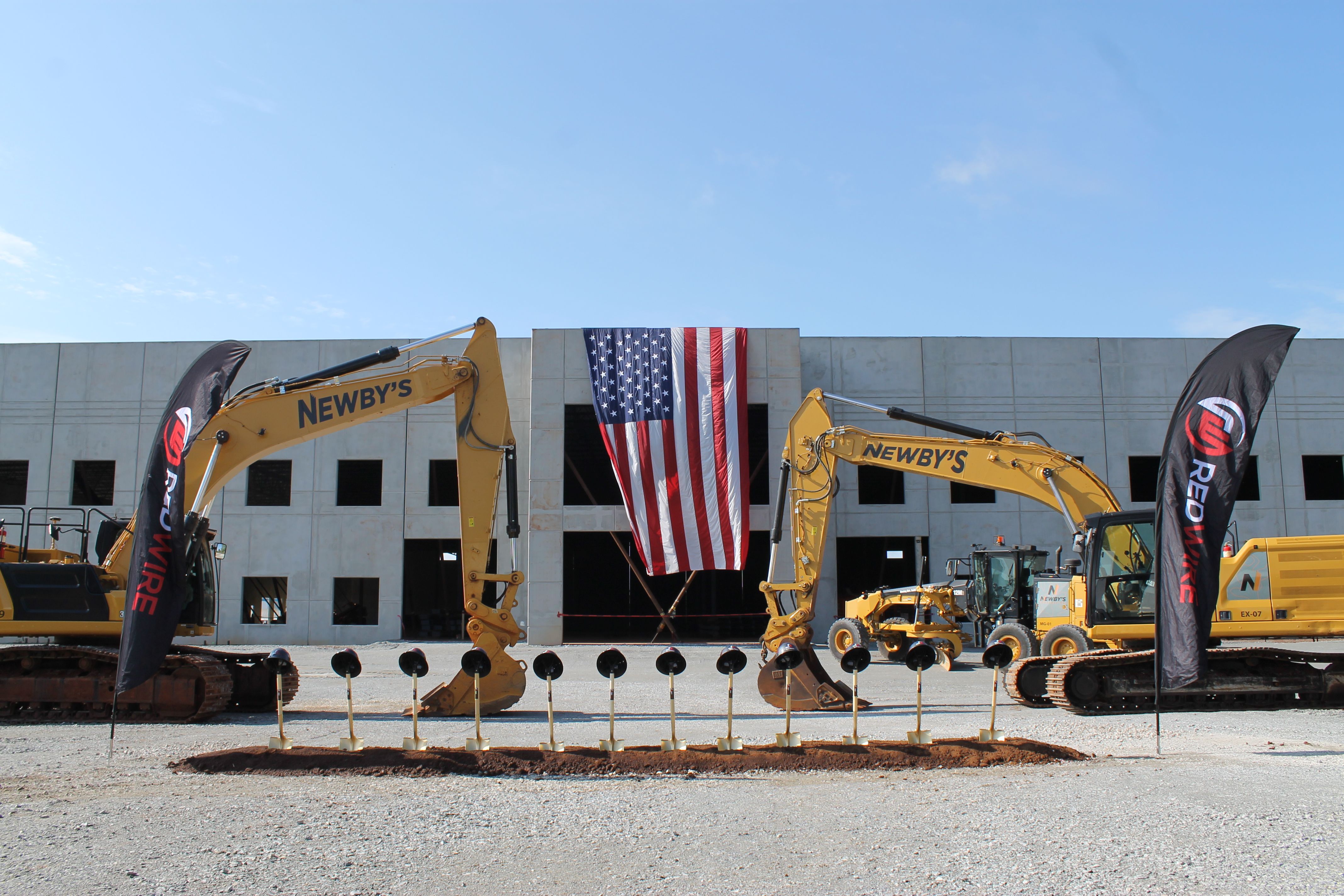 Construction site with yellow excavators in front of a gray building. A large American flag hangs between the center windows, flanked by black banners, under a clear blue sky.