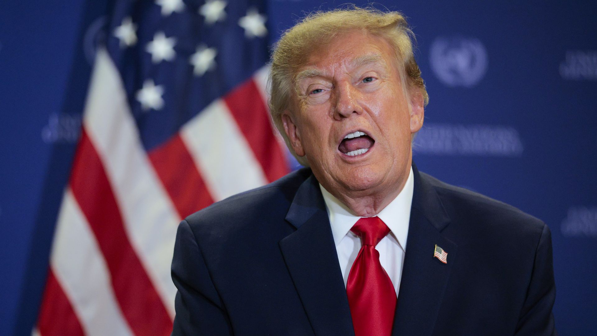 Close-up of a man with light blond hair in a dark suit and red tie, speaking with mouth open, against an American flag and a blue backdrop.