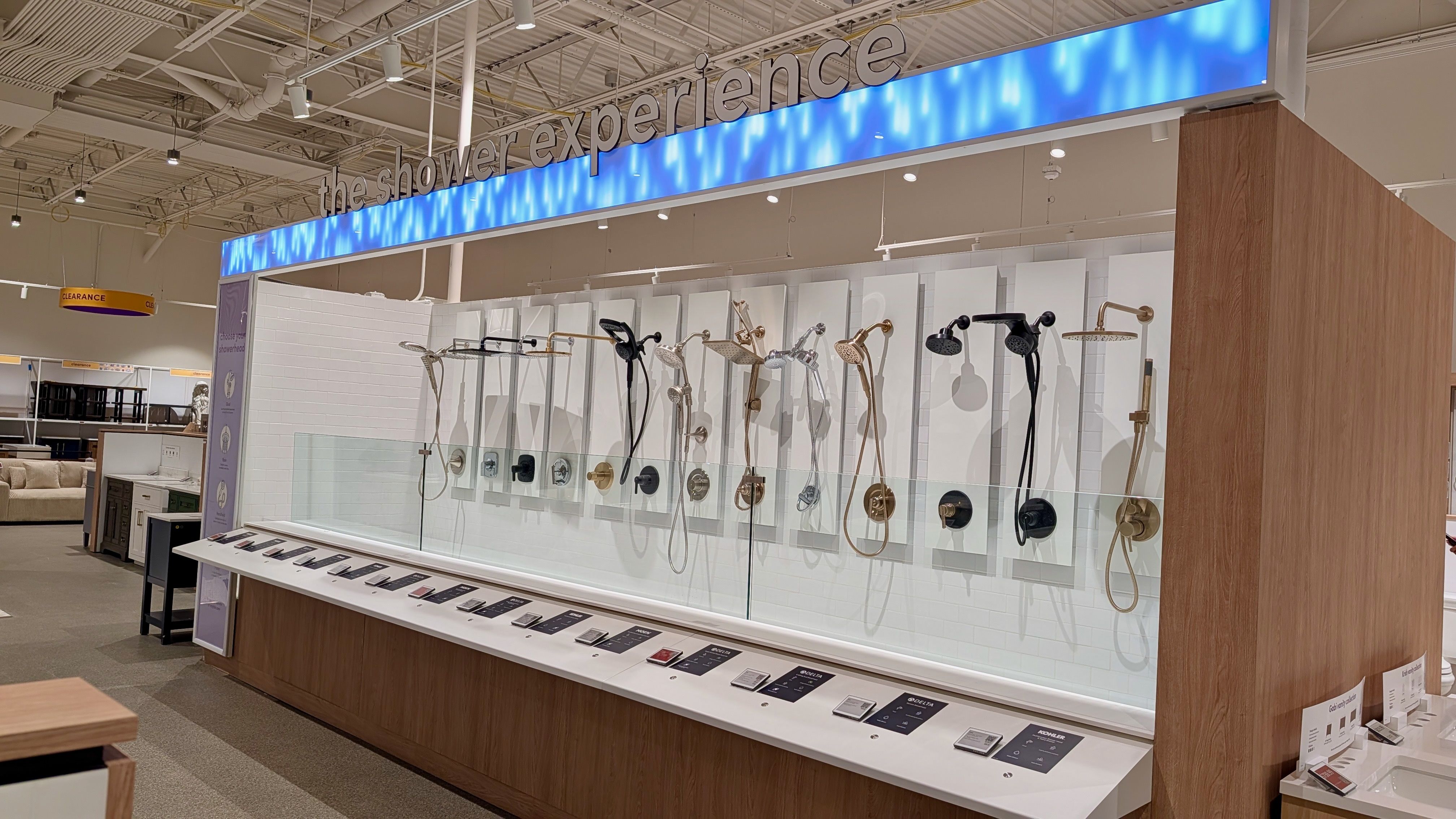 Showroom display of shower heads in bronze, chrome, and black finishes on white panels behind glass; a blue-lit sign reading the shower experience overhead; wood counter with product cards.