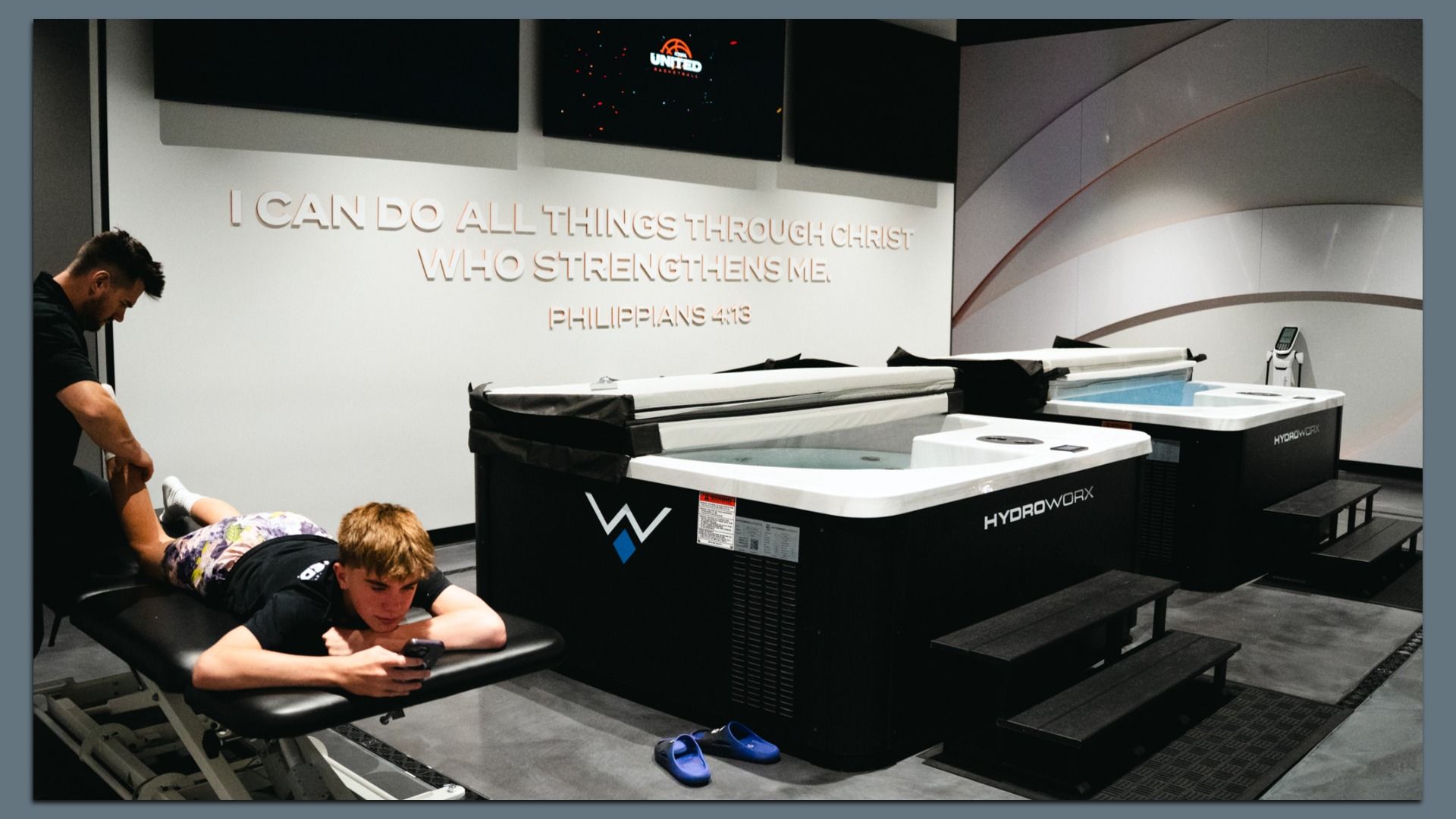 A young man lies face down on a black massage table using a phone while another person stretches his leg. Two black and white HydroWorx therapy pools are nearby. A wall displays Philippians 4:13 in gold letters.