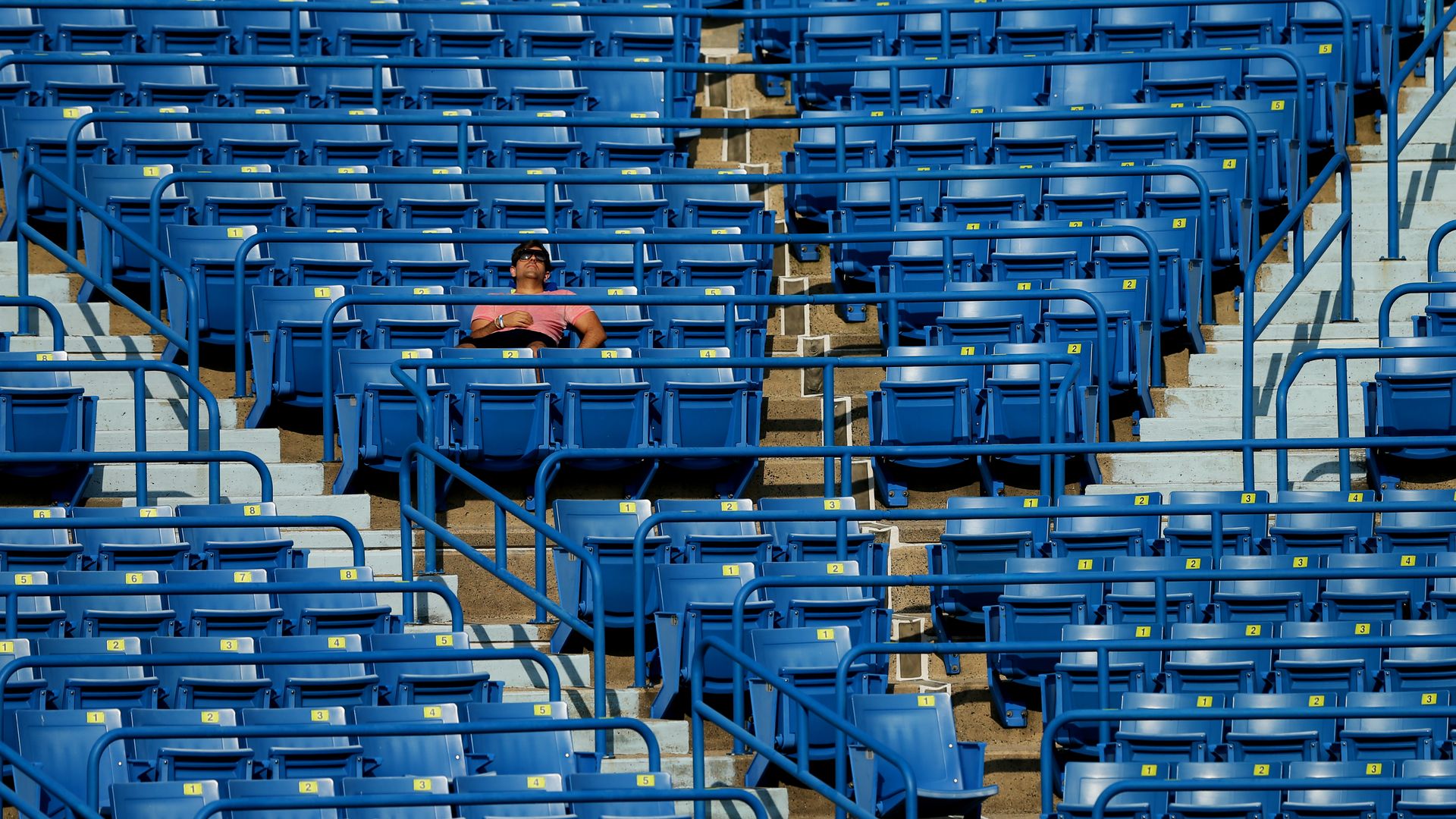 A man catches some Z's during a tennis match, surrounded by empty stadium seats.