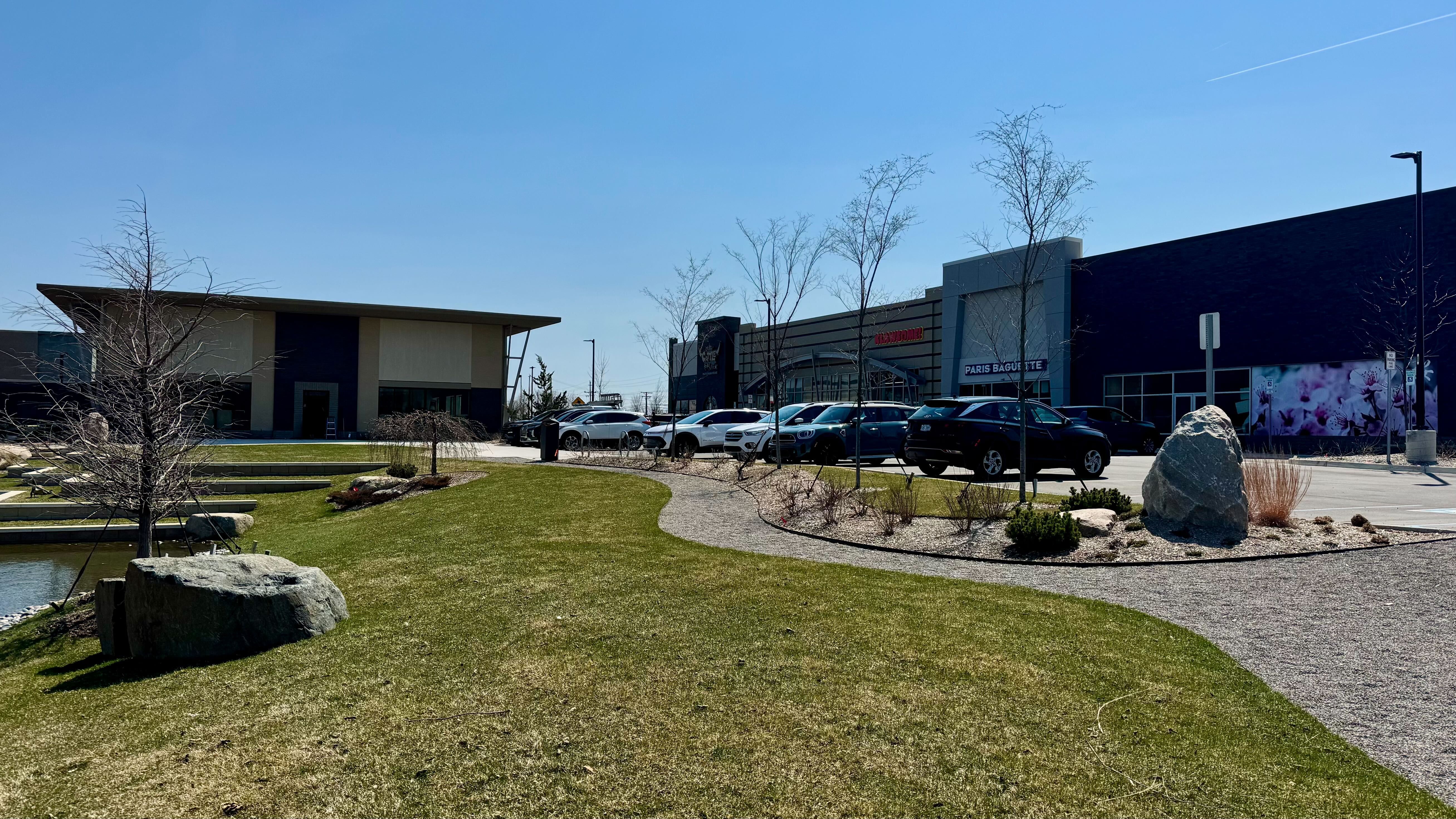 Daytime view of a modern shopping center: blue sky, cars parked along the curb, curved gravel path through green lawn with rocks and a pond, leafless trees, and a Paris Baguette storefront with a purple mural.