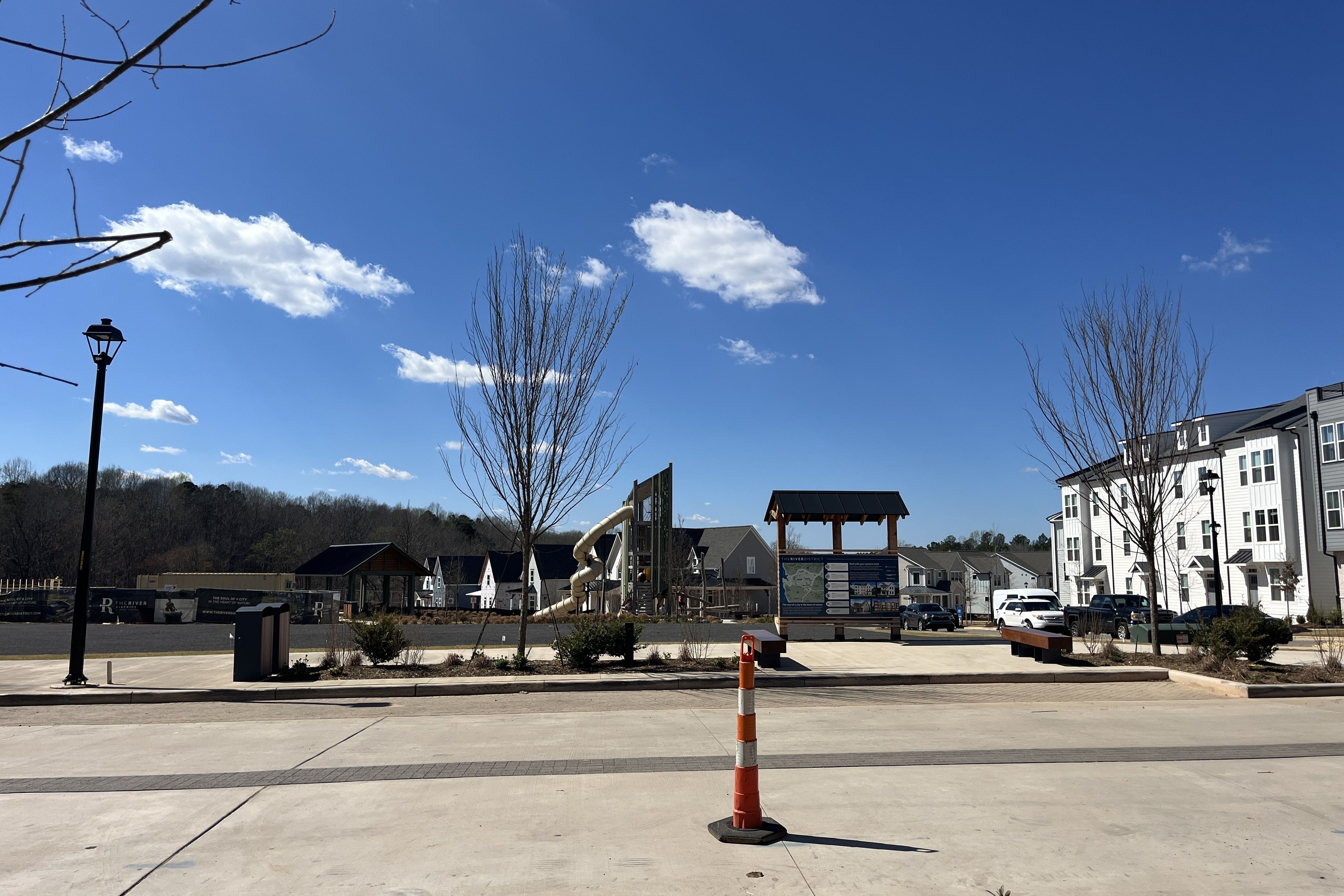 Blue-sky suburban plaza with a small playground and an info board, a twisting slide, bare trees, and white modern apartments to the right; street lamp on the left and an orange cone in foreground.