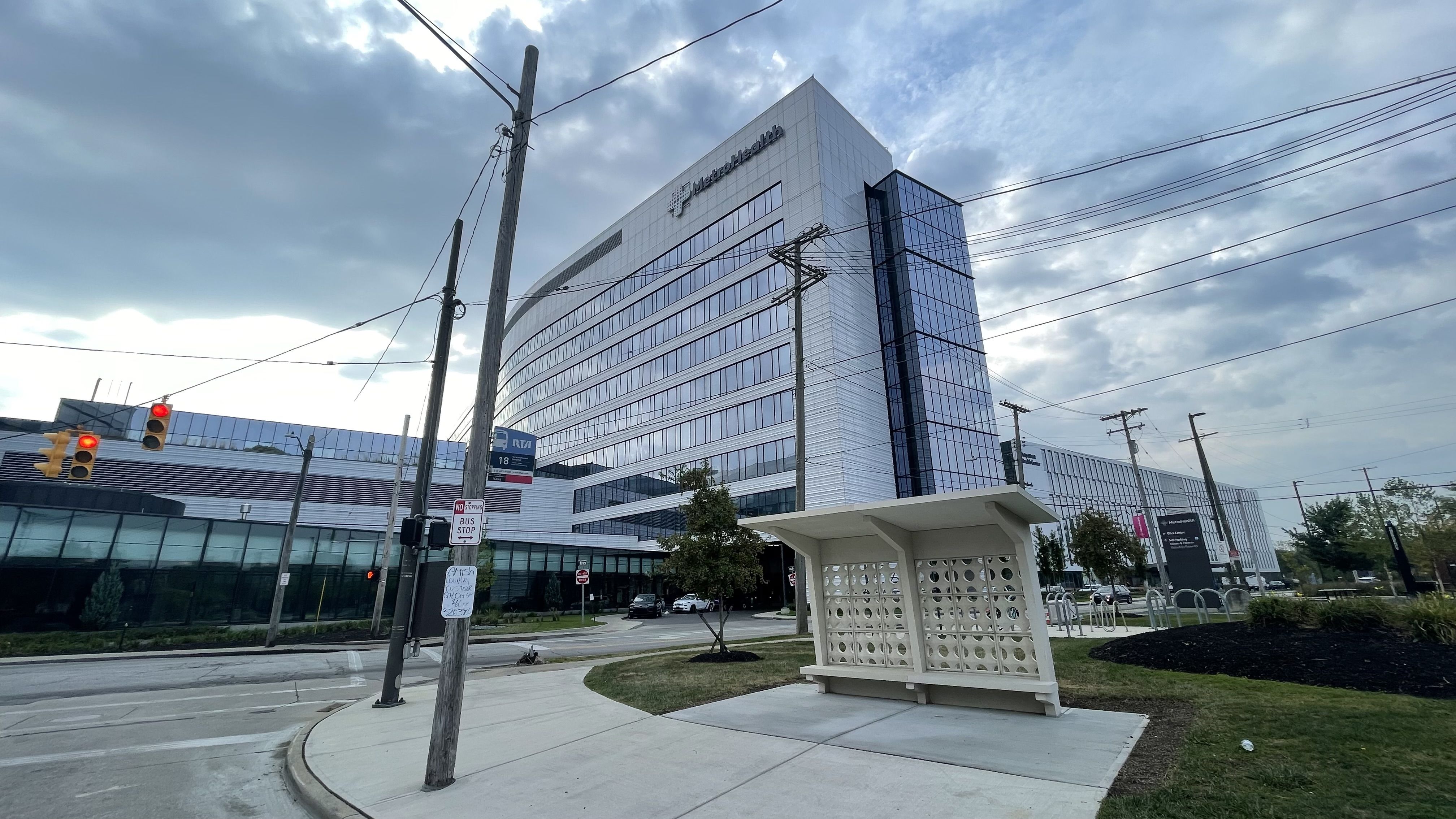 Modern multi-story MetroHealth building with reflective windows under a cloudy sky, a bus stop shelter in the foreground, and traffic lights showing red at an intersection.