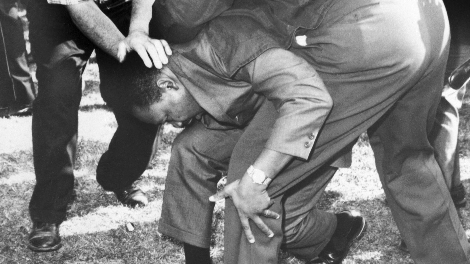Black-and-white photo of Martin Luther King kneeling with head bowed, supported by a person with a hand on his head; another man in a plaid shirt stands nearby on a grassy field.