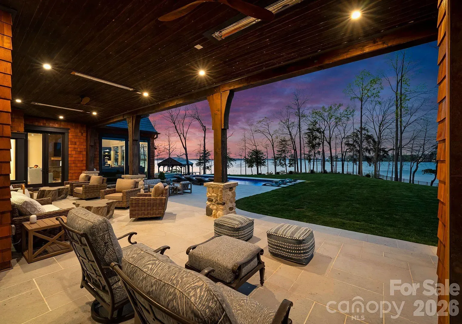 Covered outdoor seating area with wicker chairs and ottomans, stone columns, and a dark wood ceiling. Pool, green lawn, leafless trees, and a sunset over a lake in the distance.