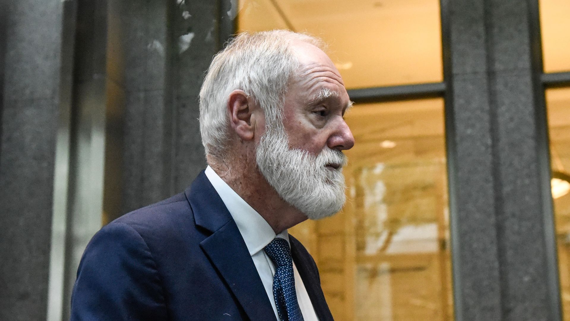 A man with a white heard looks straight ahead, slightly down, on a sidewalk
