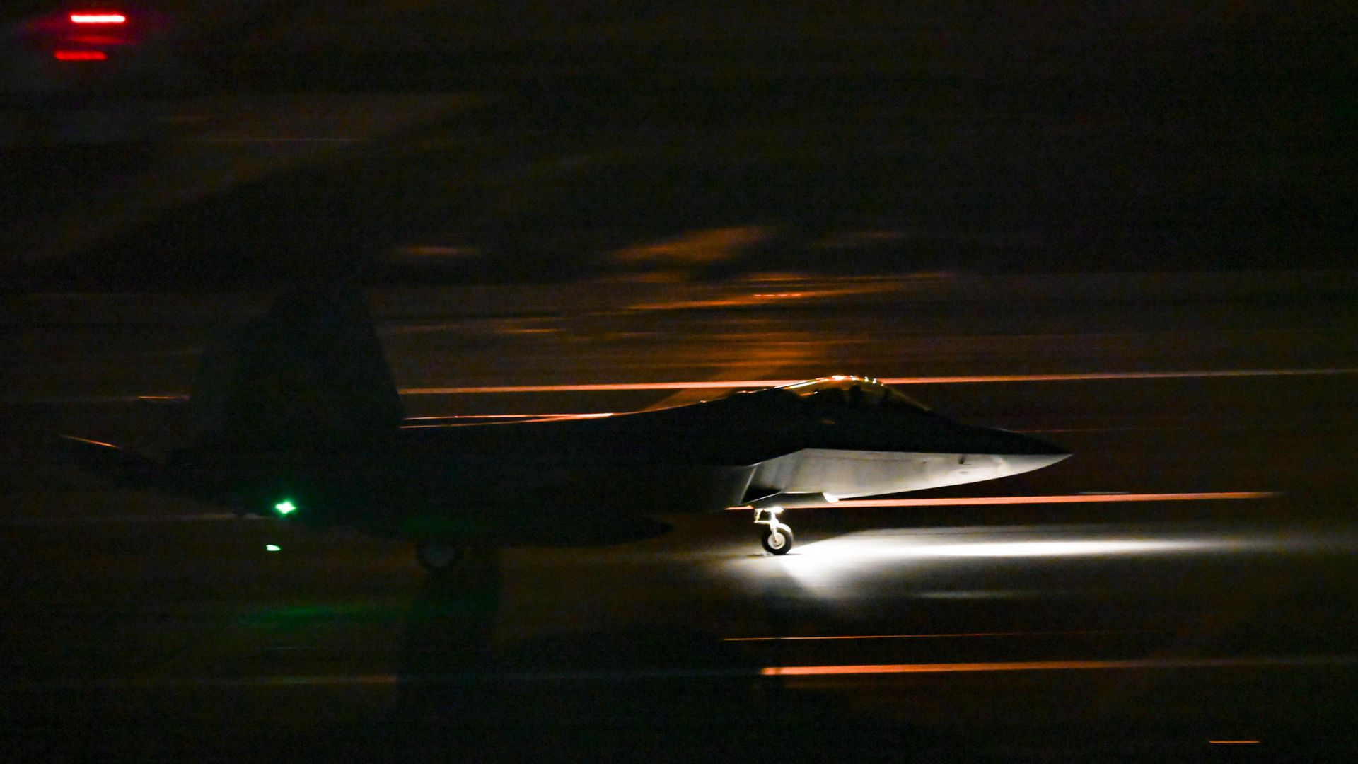 An F-22 takes off in the dark. Its nose is illuminated by lights near the front. Its tail is back lit.