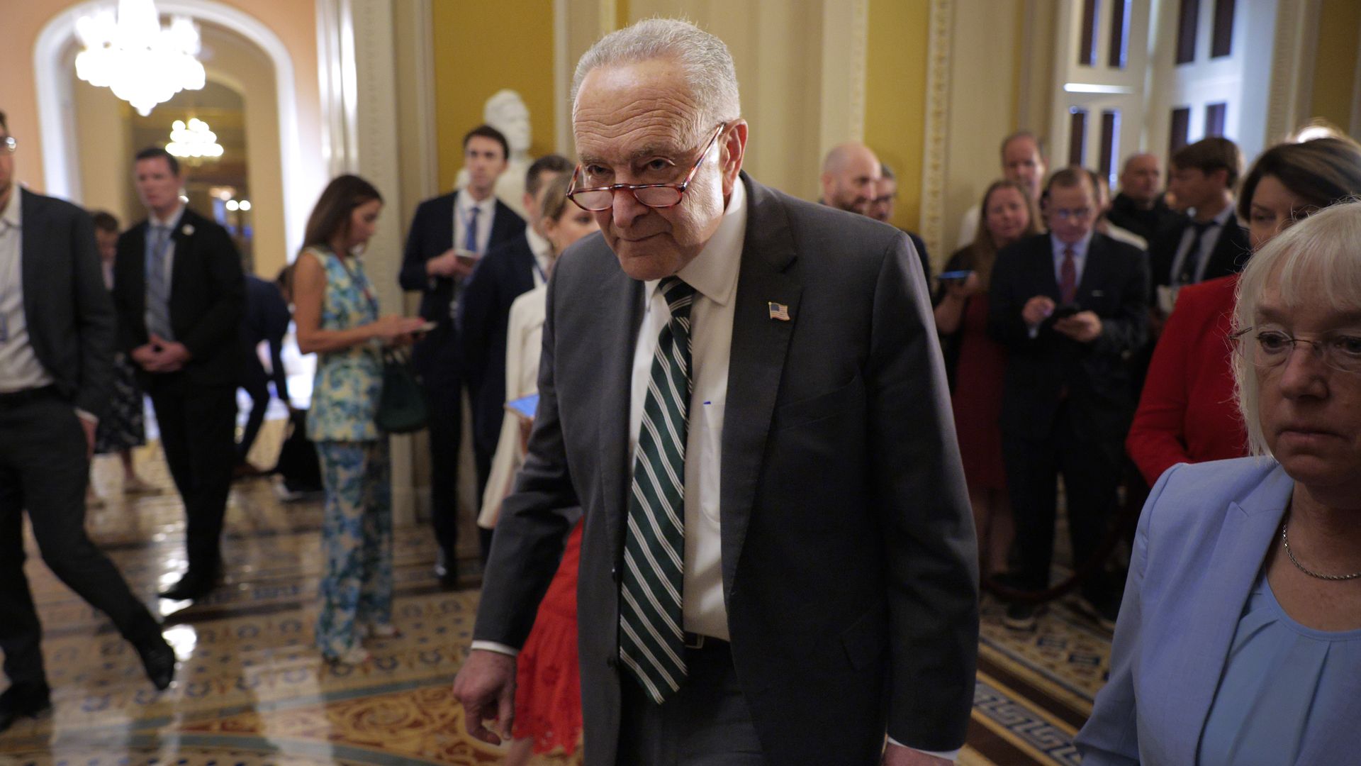 An older man wearing glasses, a dark suit, white shirt, and green-and-white striped tie walks through a decorative hallway with a crowd of people behind him, some using phones.