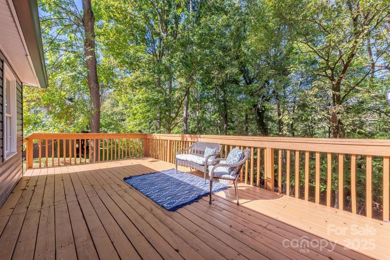Sunny wooden deck with railings, featuring a blue patterned rug, metal loveseat and chair with cushions, surrounded by green trees and part of a house on the left.