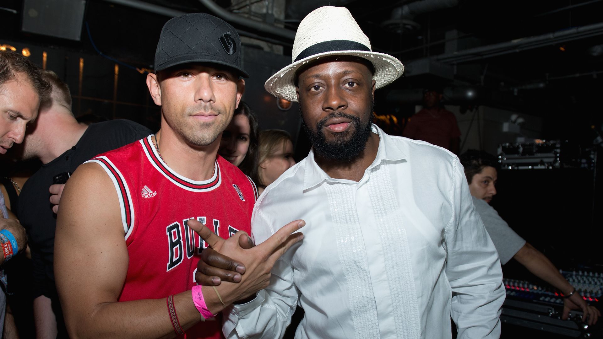 Two men posing for a photo indoors; one wears a black cap and red Chicago Bulls jersey, the other a white hat and white shirt, both making hand gestures.
