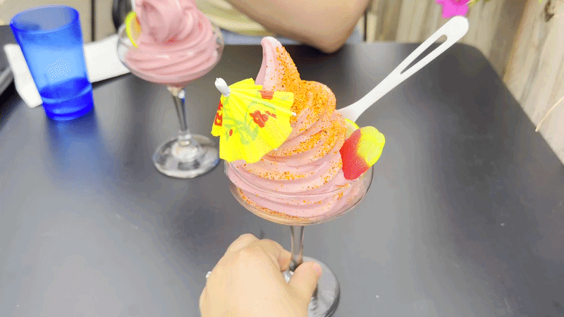 A pink soft-serve sundae swirled in a tall glass, garnished with a yellow paper umbrella and fruit candy, held by a hand at a dark table; a blue cup and another pink dessert on the background.