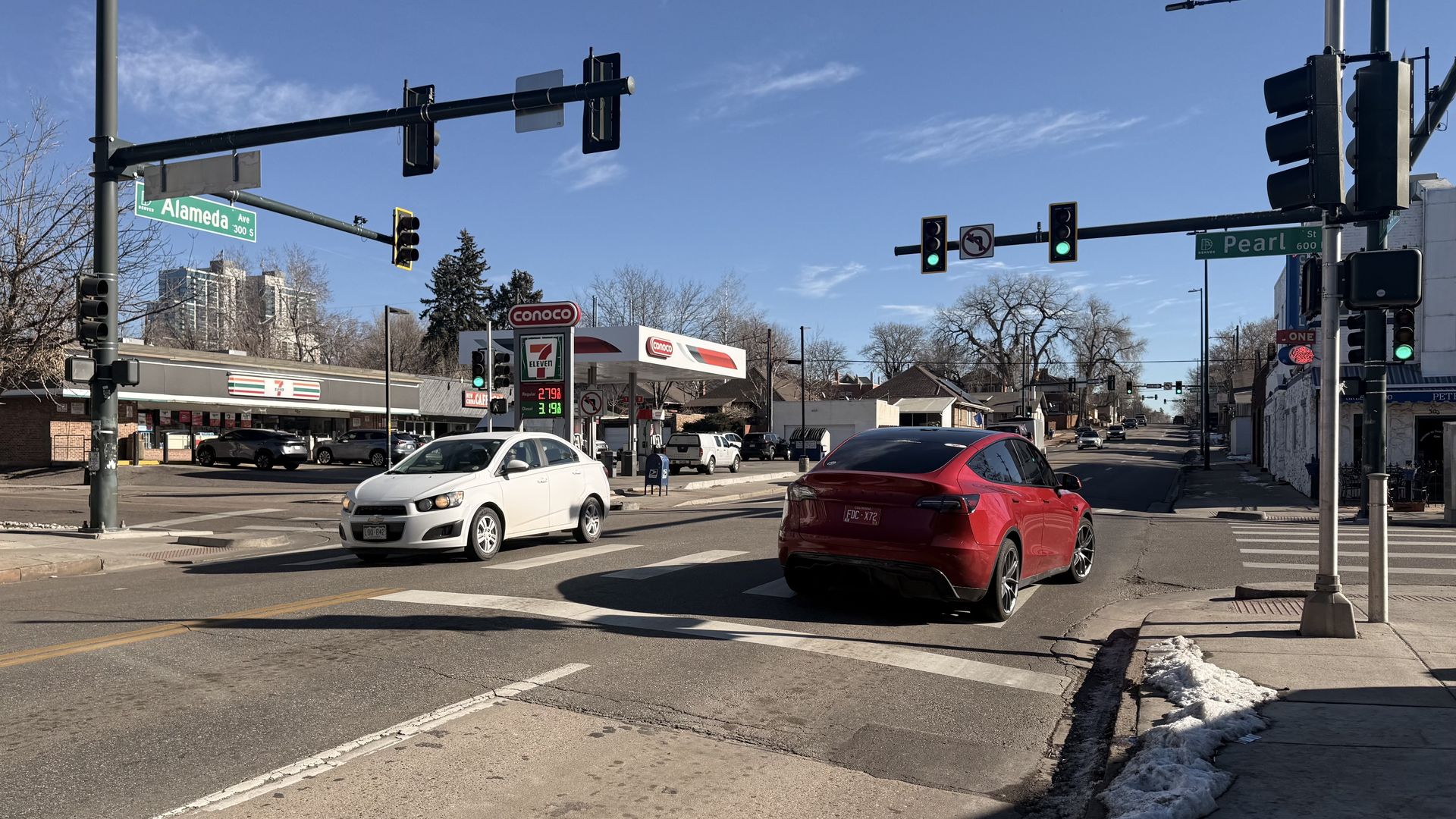Sunny street intersection with traffic lights showing green. A red car and a white car are stopped. A 7-Eleven and Conoco gas station are in the background. Street signs read Alameda Ave and Pearl St.