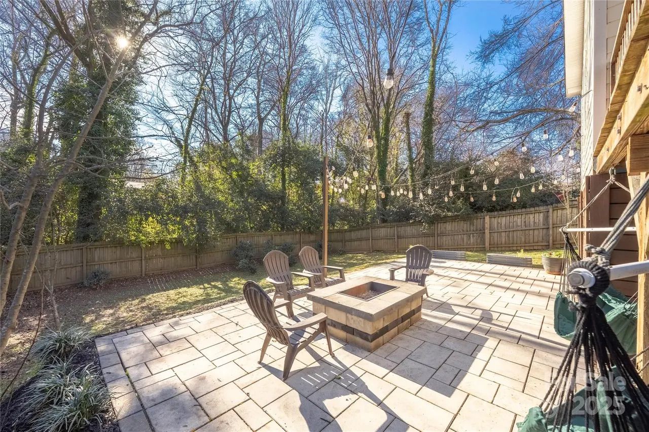 Sunny backyard patio with stone tile flooring, a square fire pit surrounded by four plastic chairs, string lights hanging above, wooden fence, and leafless trees in the background.