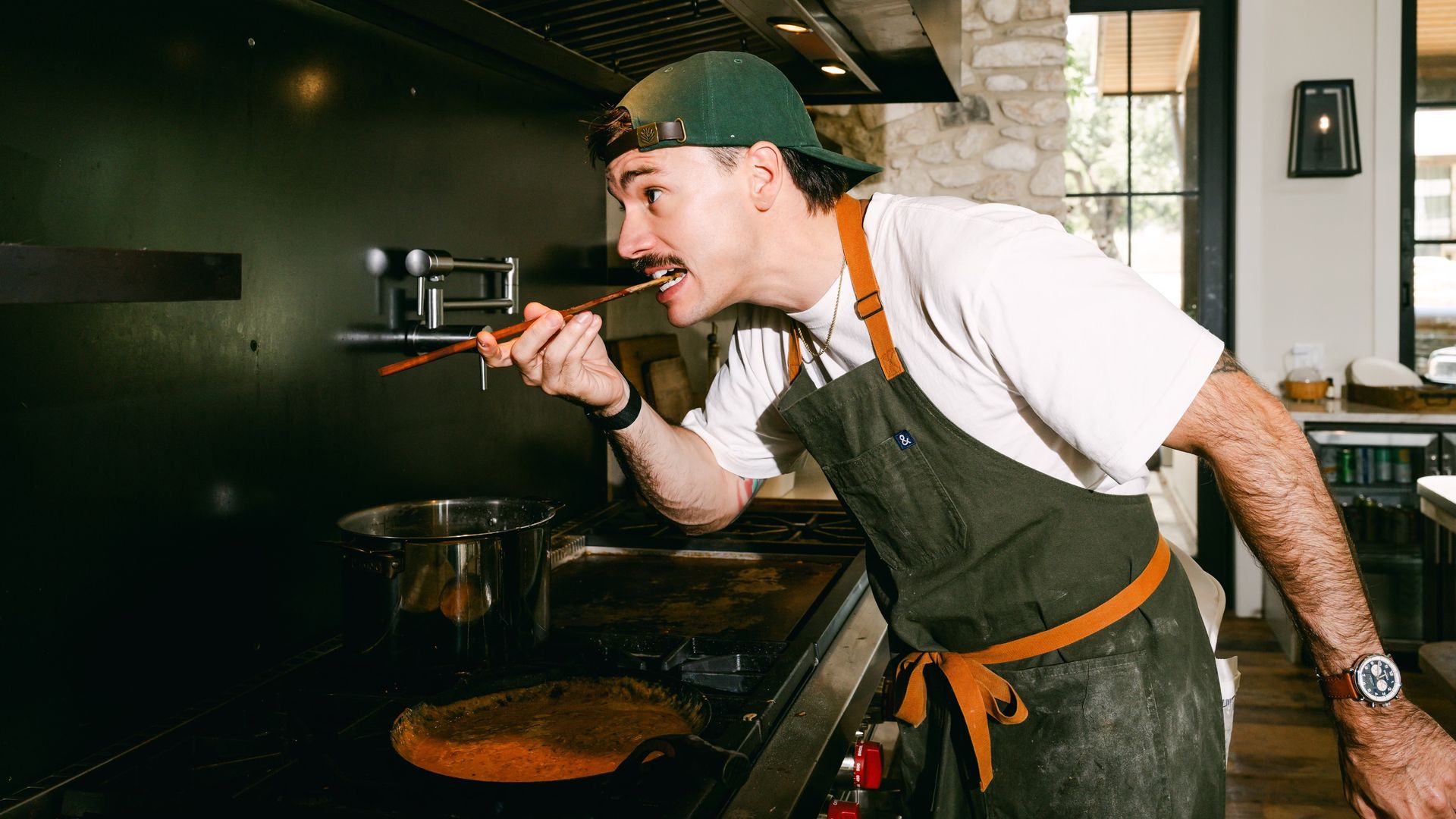 A man wearing a green cap and dark apron with orange straps tastes orange sauce from a wooden spoon at a stove in a modern kitchen with a stone wall and large windows.