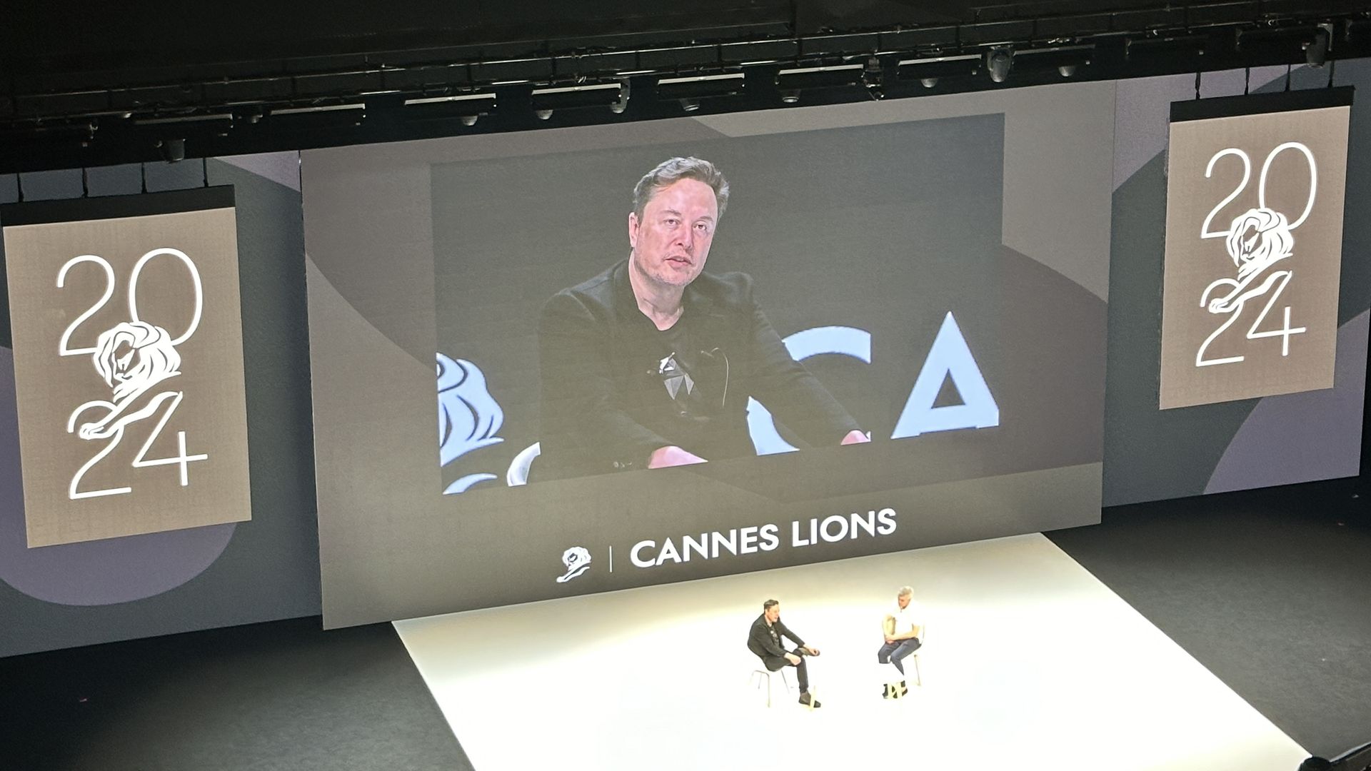 Elon Musk and Mark Read sitting in chairs on a stage with a screen in the back that says Cannes Lions