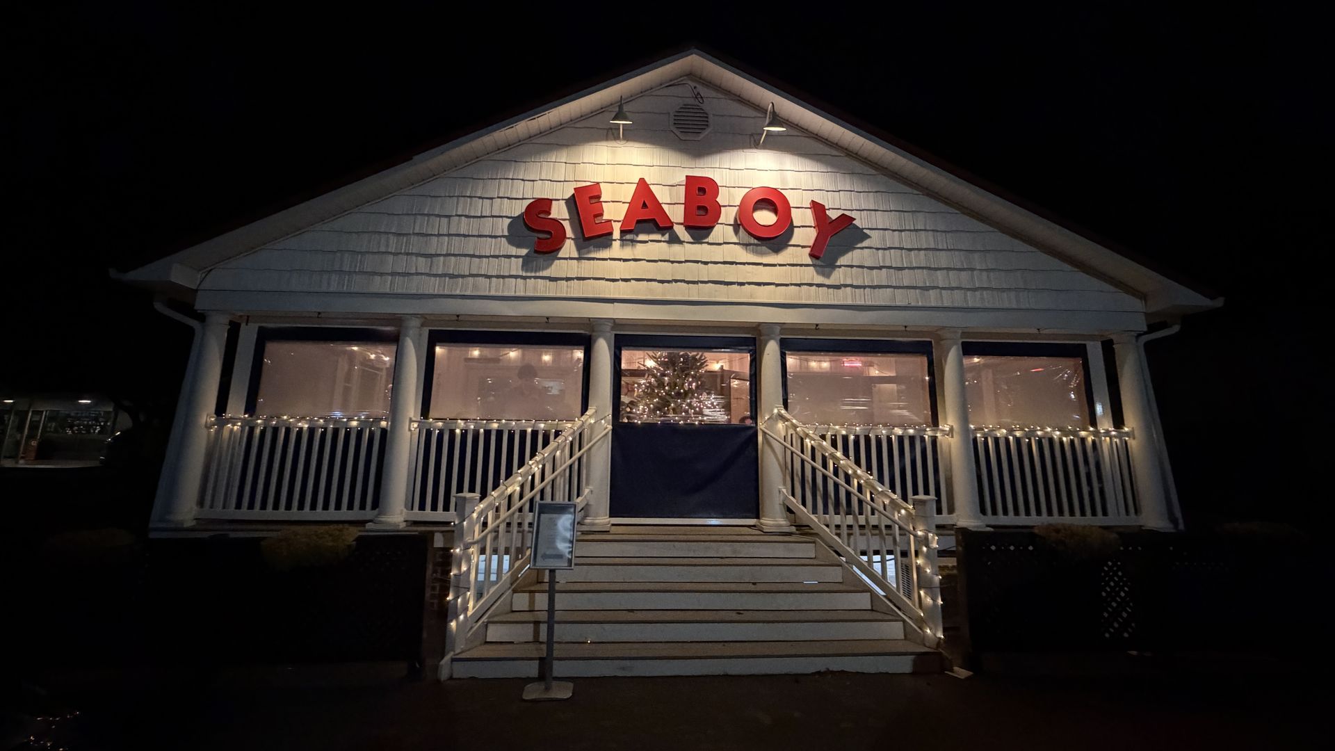 Night view of a white building with red letters spelling SEABOY, decorated with string lights on the stairs and railings, and a lit Christmas tree visible inside.