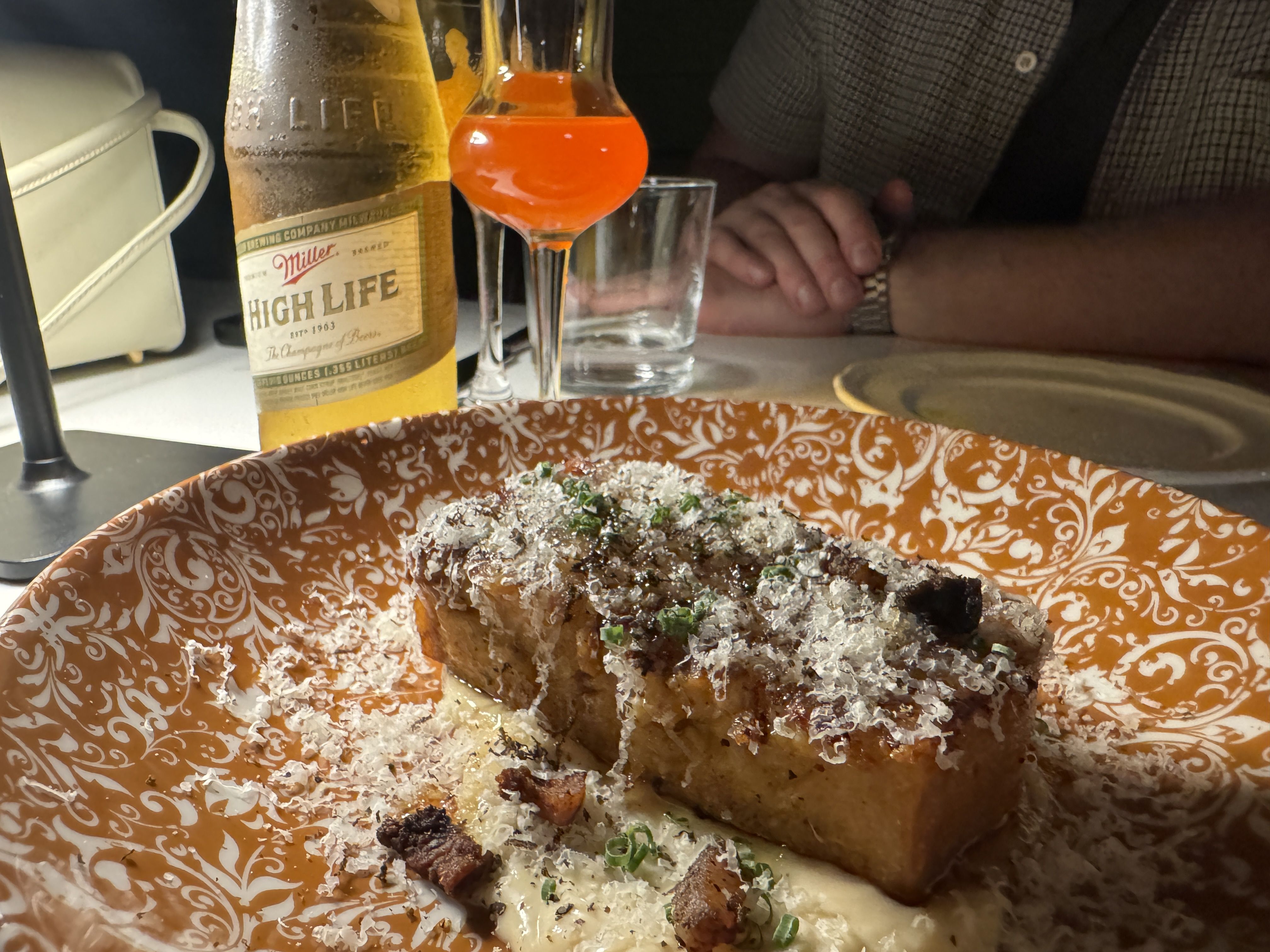 Close-up of a richly garnished dish on an orange patterned plate with grated cheese, small green herbs, and crispy bits, accompanied by a bottle of Miller High Life beer and a glass with orange liquid.