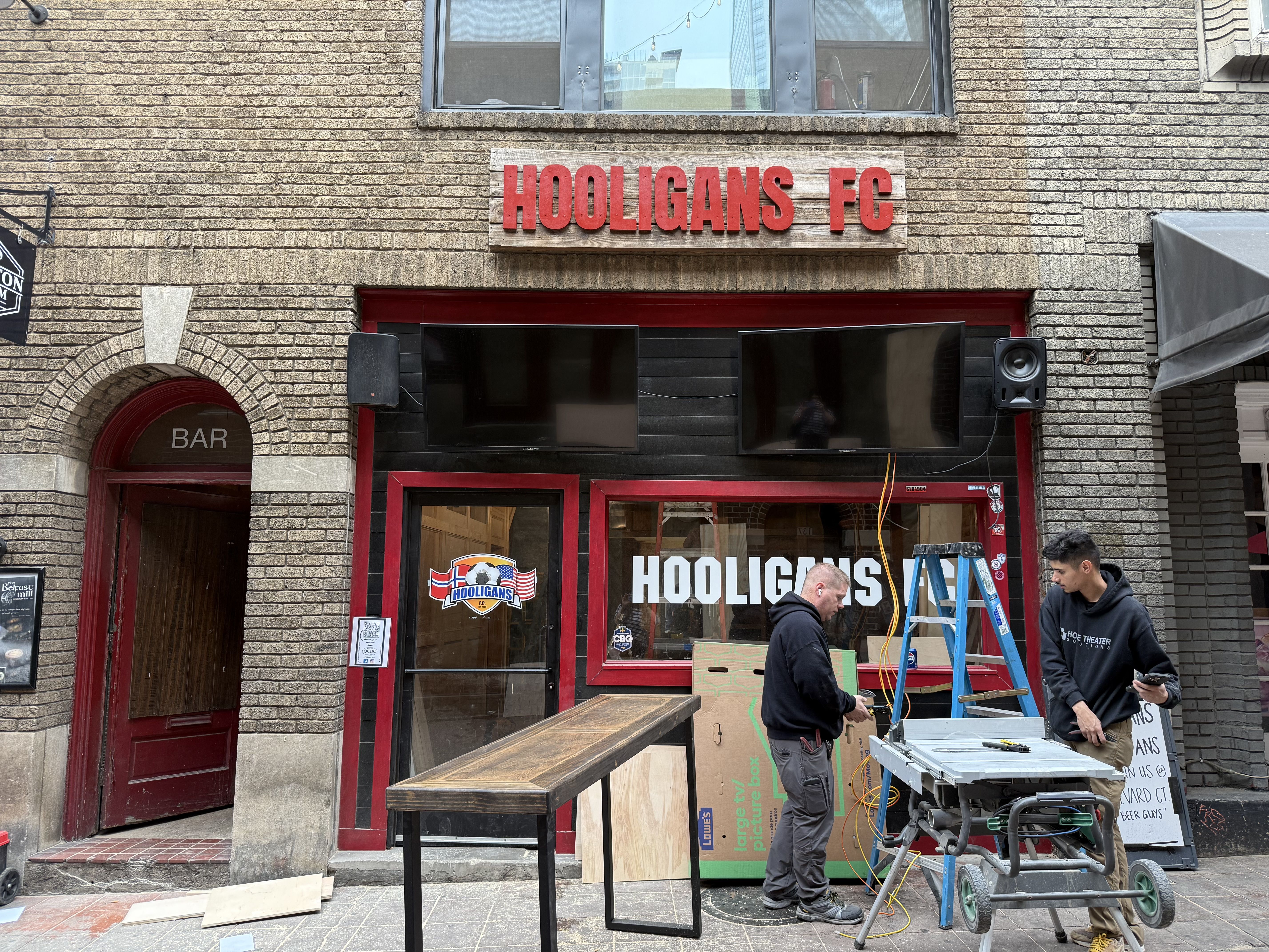 Two men working outside Hooligans FC, a bar with red and black entrance, wooden table, blue ladder, and tools on sidewalk in front of brick building.