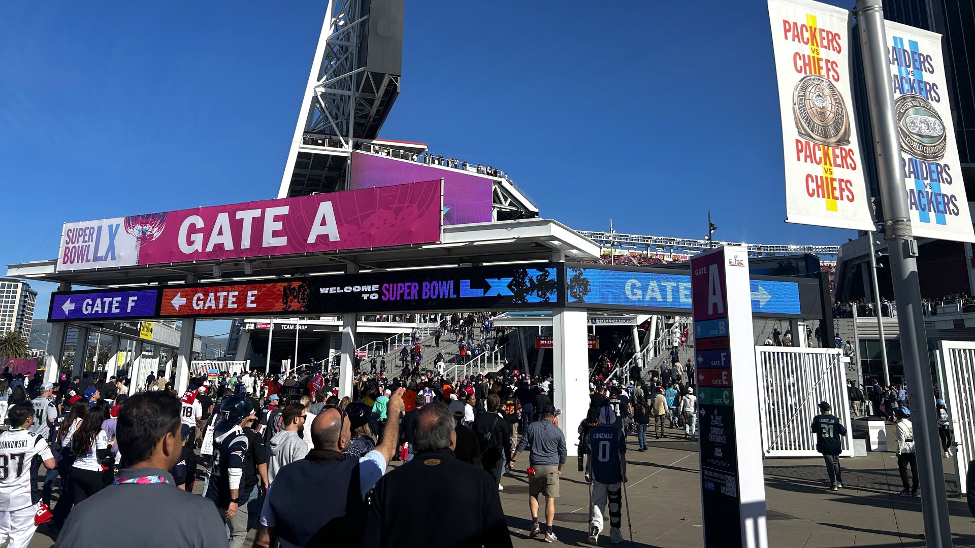 Crowd of fans entering Gate A at a stadium for Super Bowl LX on a clear sunny day, with purple signage and banners showing Packers vs Chiefs and Raiders vs Packers games.