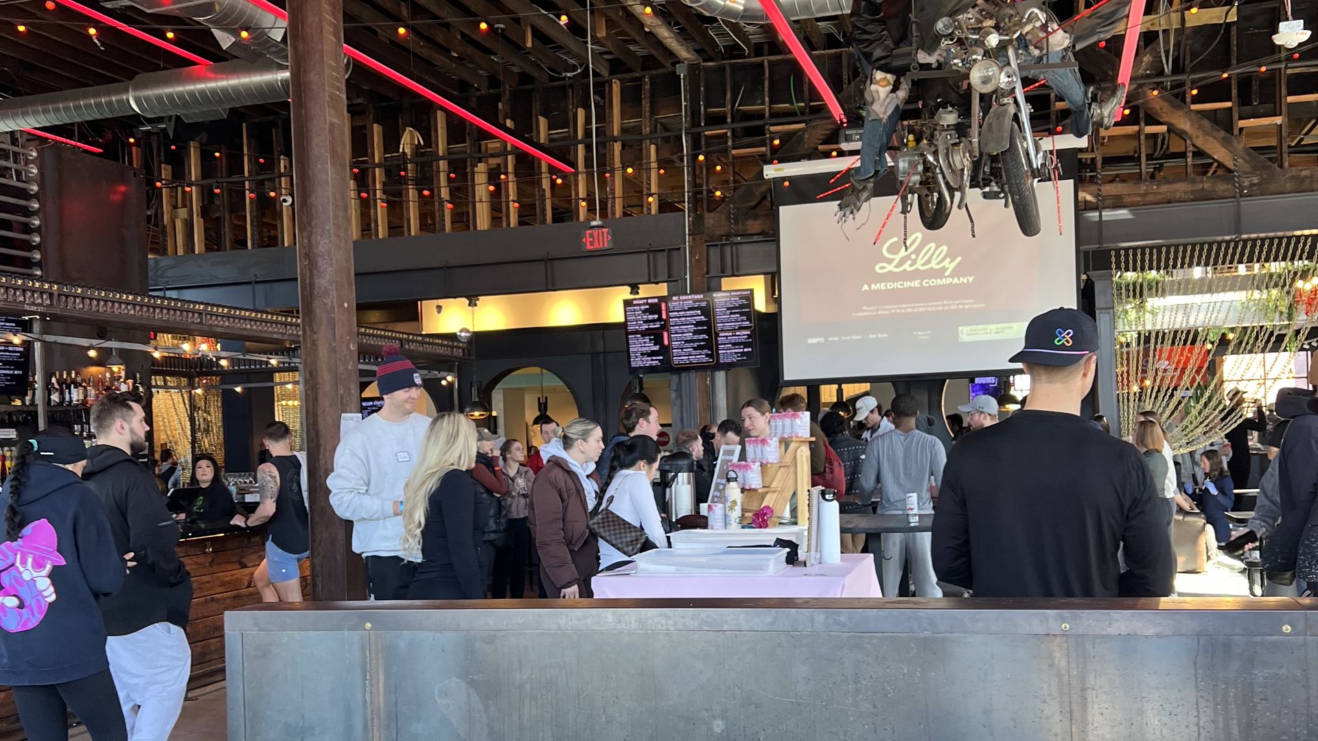Crowded indoor bar with wooden barrels, exposed ceiling beams, red string lights, and a skeleton mannequin on a motorcycle hanging from the ceiling. People are gathered around the counter.