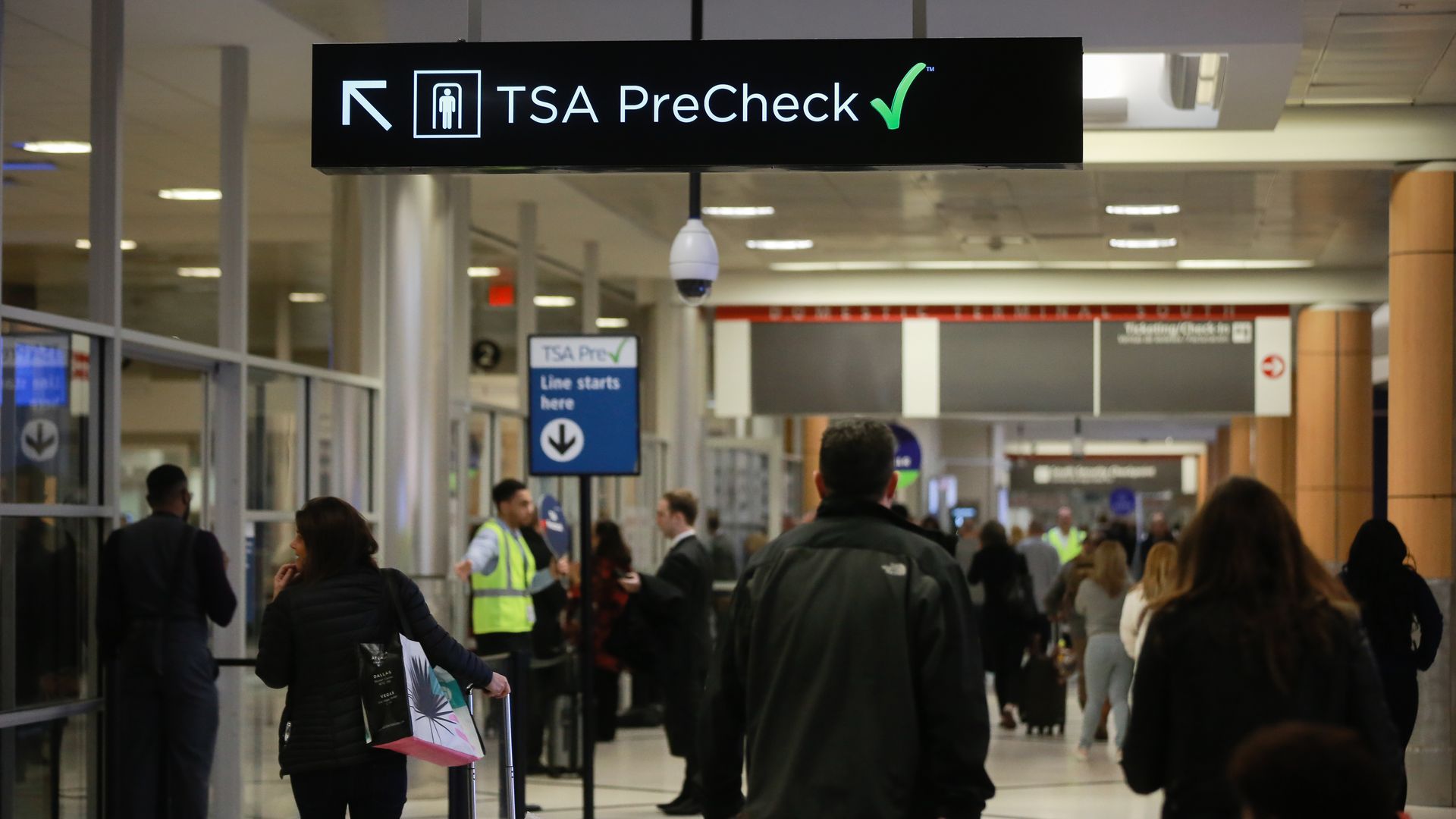 Photo of a sign that says "TSA PreCheck" in a hallway with passengers carrying suitcases