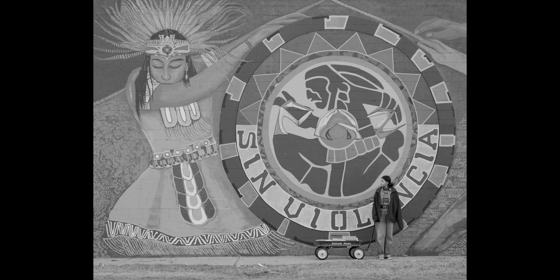 A still from a movie. A person stands in front of a mural. 