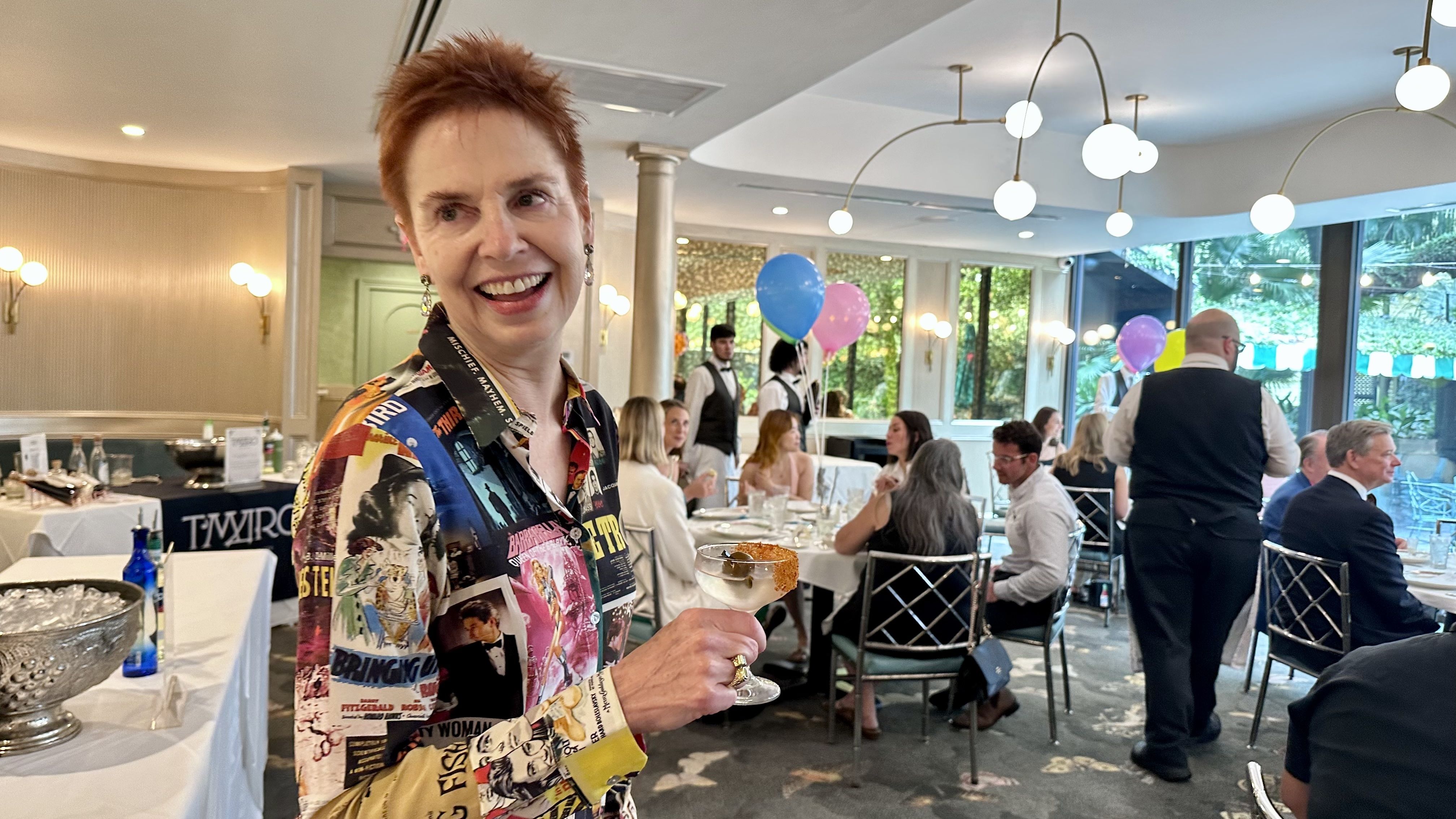 Smiling woman with short red hair wearing a colorful, graphic print shirt holds a cocktail glass with an olive and orange garnish in a bright restaurant filled with people, balloons and waitstaff.