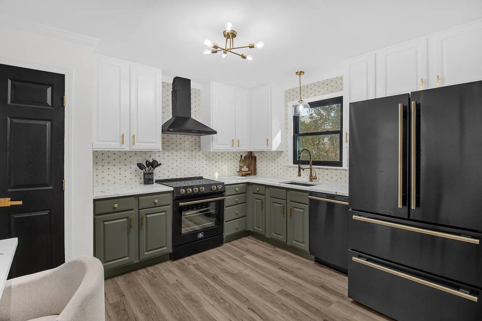 Modern kitchen with black appliances, white upper cabinets, green lower cabinets, brass hardware, wood-look flooring, patterned backsplash, and large window over sink.