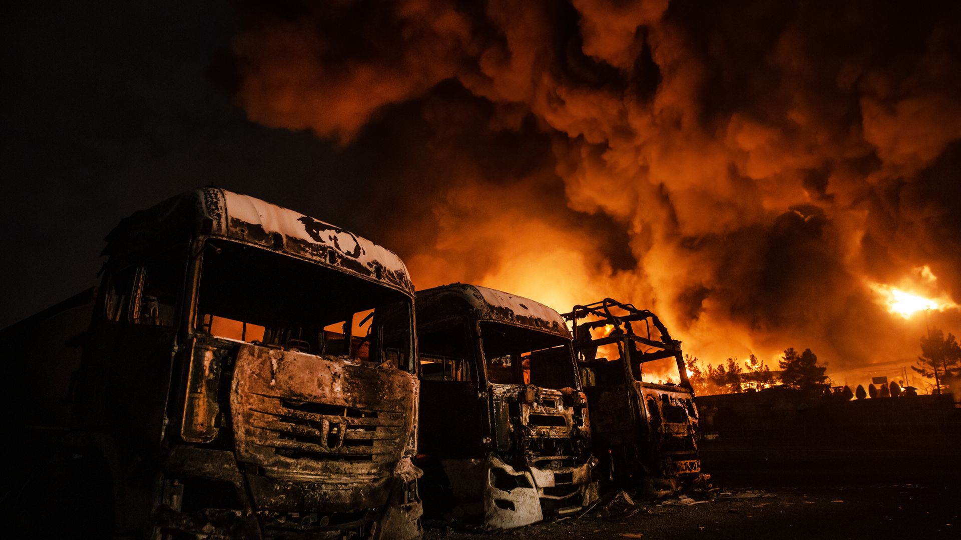 Three vehicles in various states of damage are parked against the backdrop of a flaming orange and black sky in Iran. 