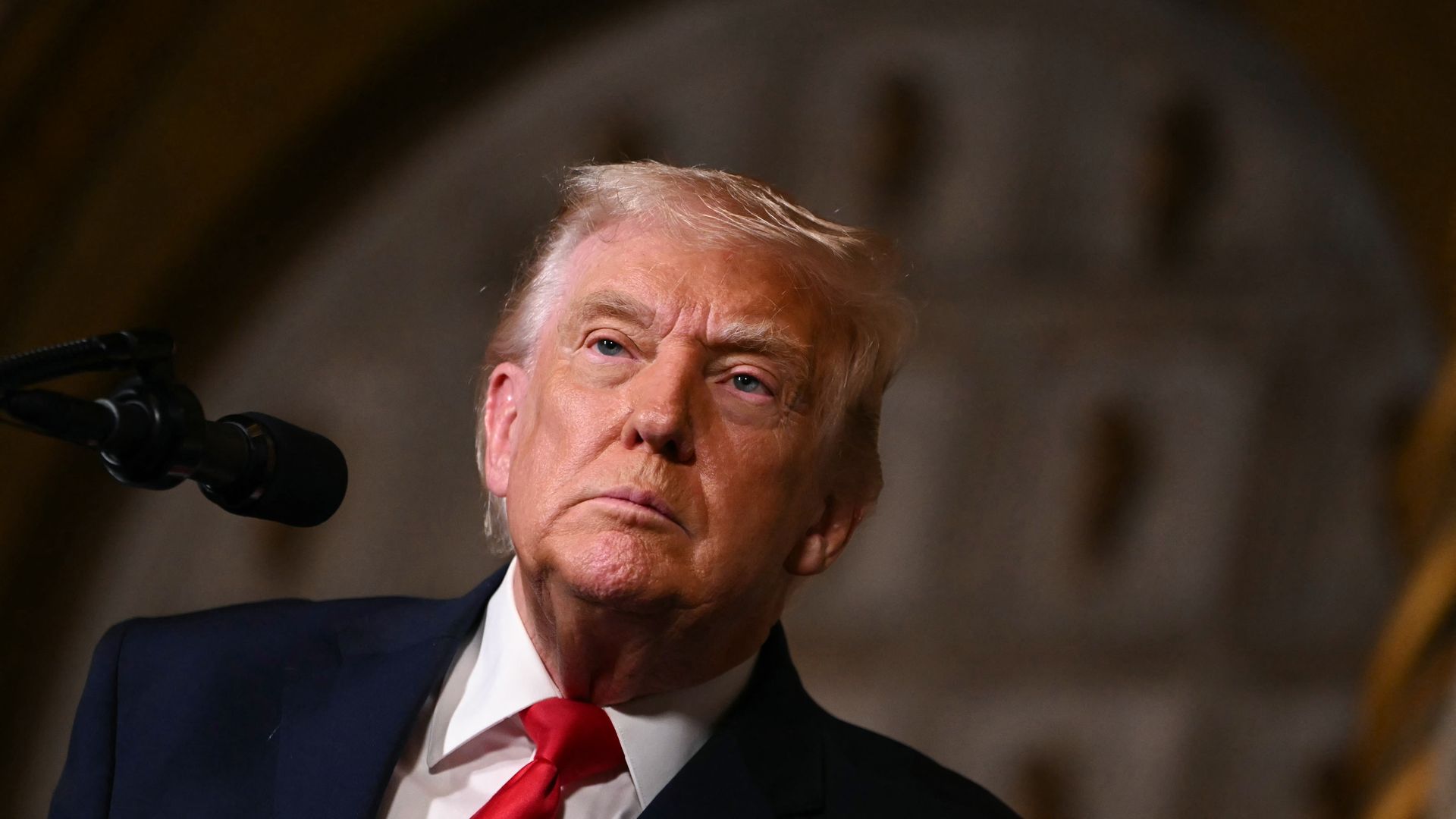President Trump — wearing a dark suit, a white collared shirt and a red tie — looks on as he stands behind a microphone.